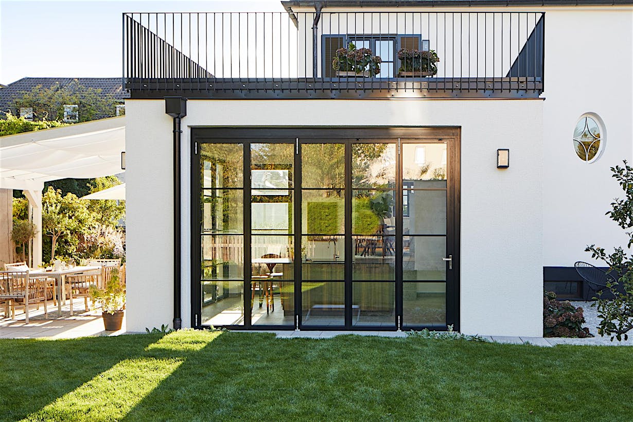 Modern house exterior with accordion glass walls, large black-framed doors, a balcony above with potted plants, and a grassy lawn in the foreground.