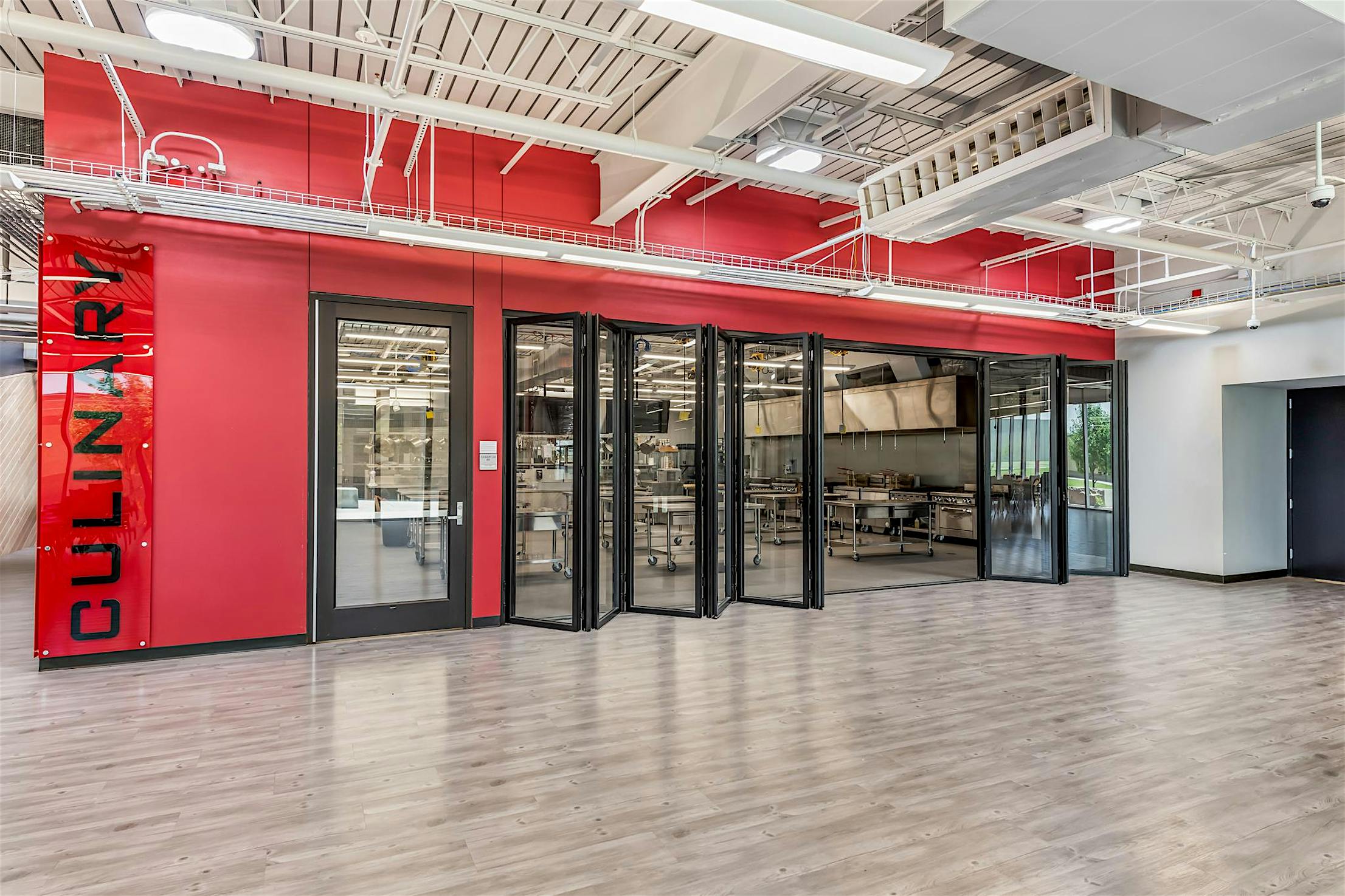 Modern culinary classroom with red walls and large glass folding walls, revealing a kitchen with stainless steel appliances and cooking stations inside.