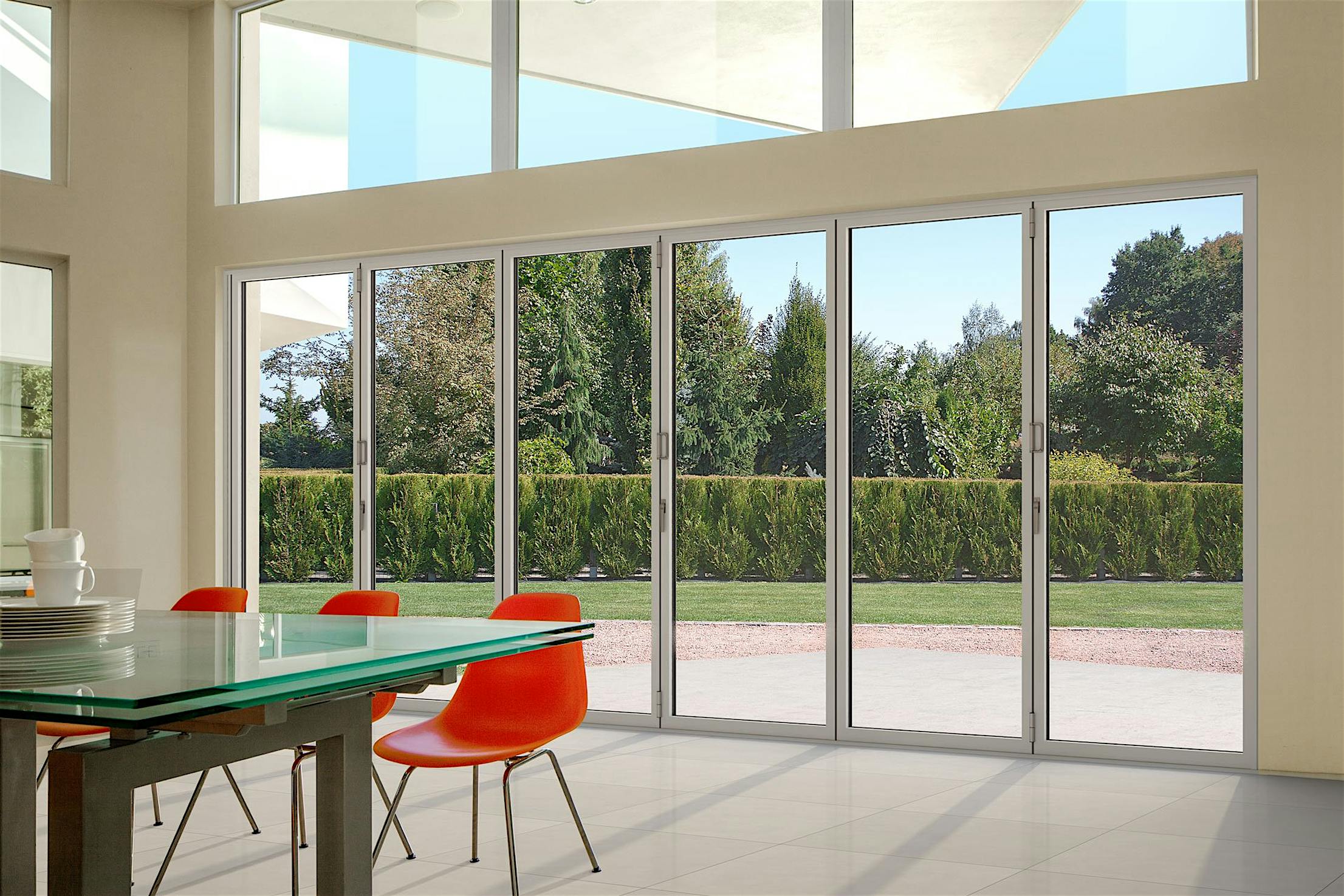 Stylish dining area with orange chairs and a glass table, overlooking a green yard with trees through large interior glass doors