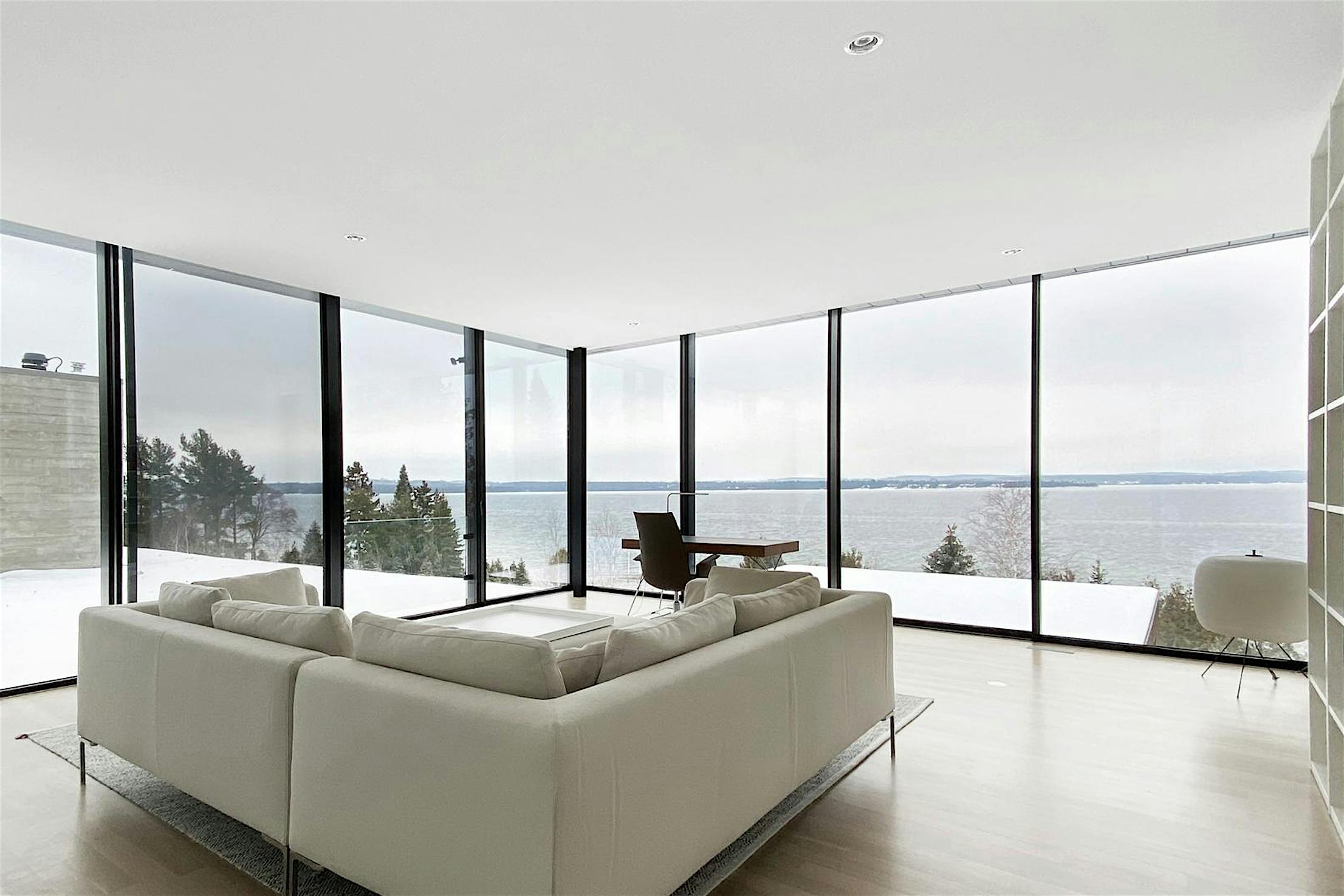 Minimalist living room with a white sofa, sleek floor-to-Ceiling sliding glass doors, serene snowy and water views, light wood flooring, and modern built-in shelving.