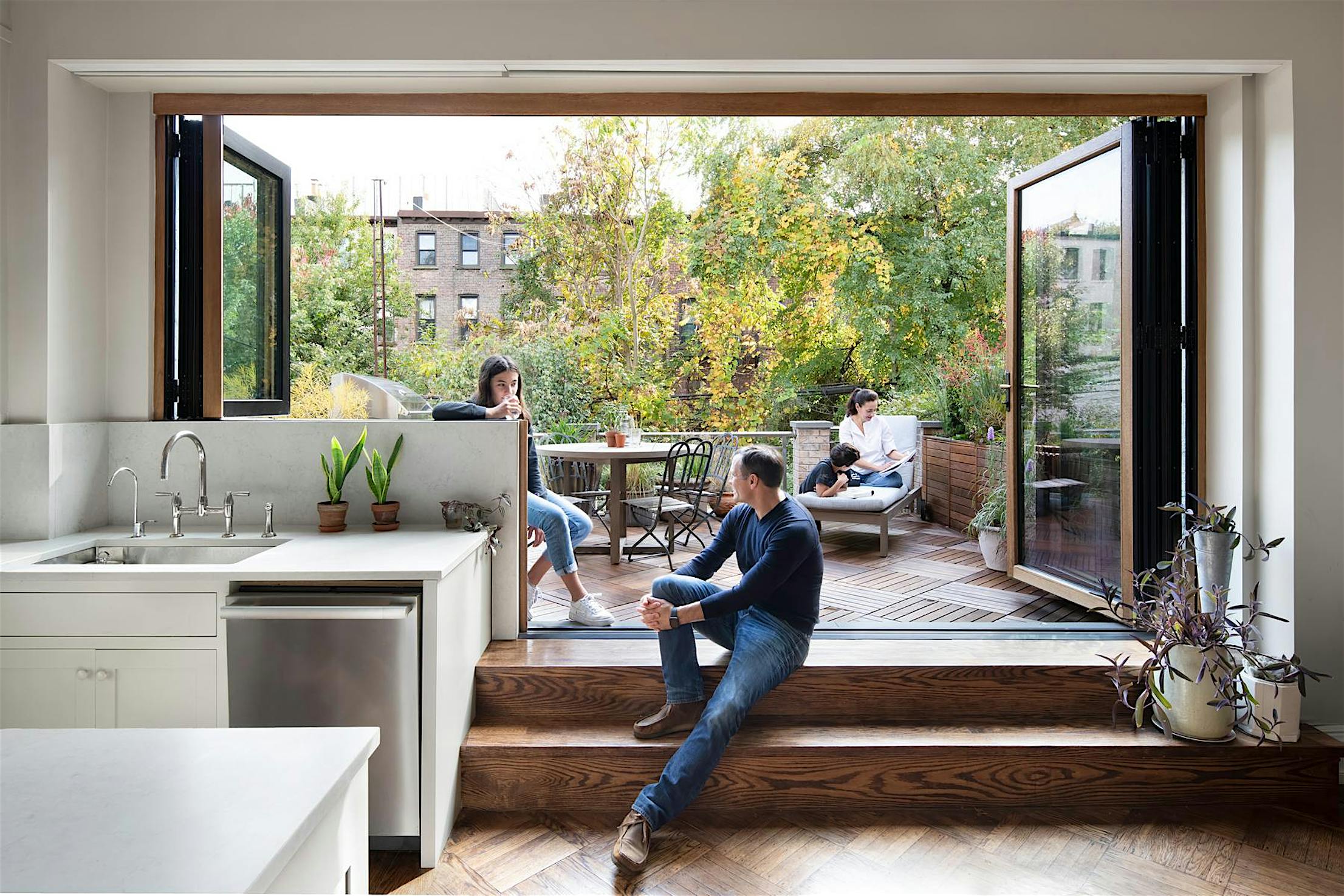 Modern kitchen with retractable glass wall that opens the kitchen to a patio. Three people relax in a modern home with a retractable glass wall that opens the kitchen to a patio; two people sit outside amid greenery, while one sits on indoor steps next to houseplants.