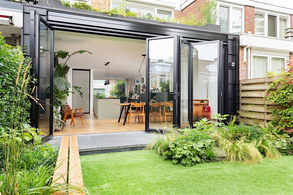 Modern home with large glass doors opening to a green garden, showing a spacious, light-filled kitchen and dining area with wooden flooring and contemporary furniture.