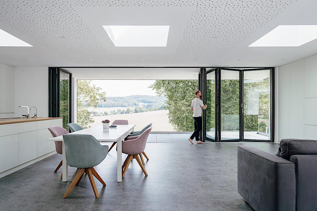 A person opens folding glass doors in a modern, open-plan living and dining area, seamlessly connecting the space to scenic views of trees and fields outside.