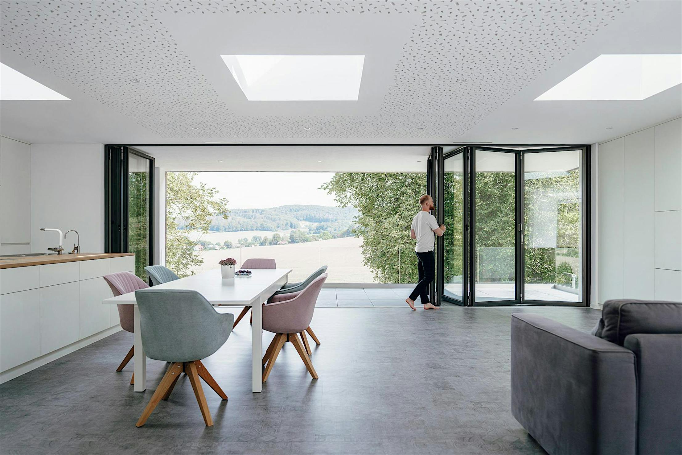 A person opens folding glass doors in a modern, open-plan living and dining area, seamlessly connecting the space to scenic views of trees and fields outside.