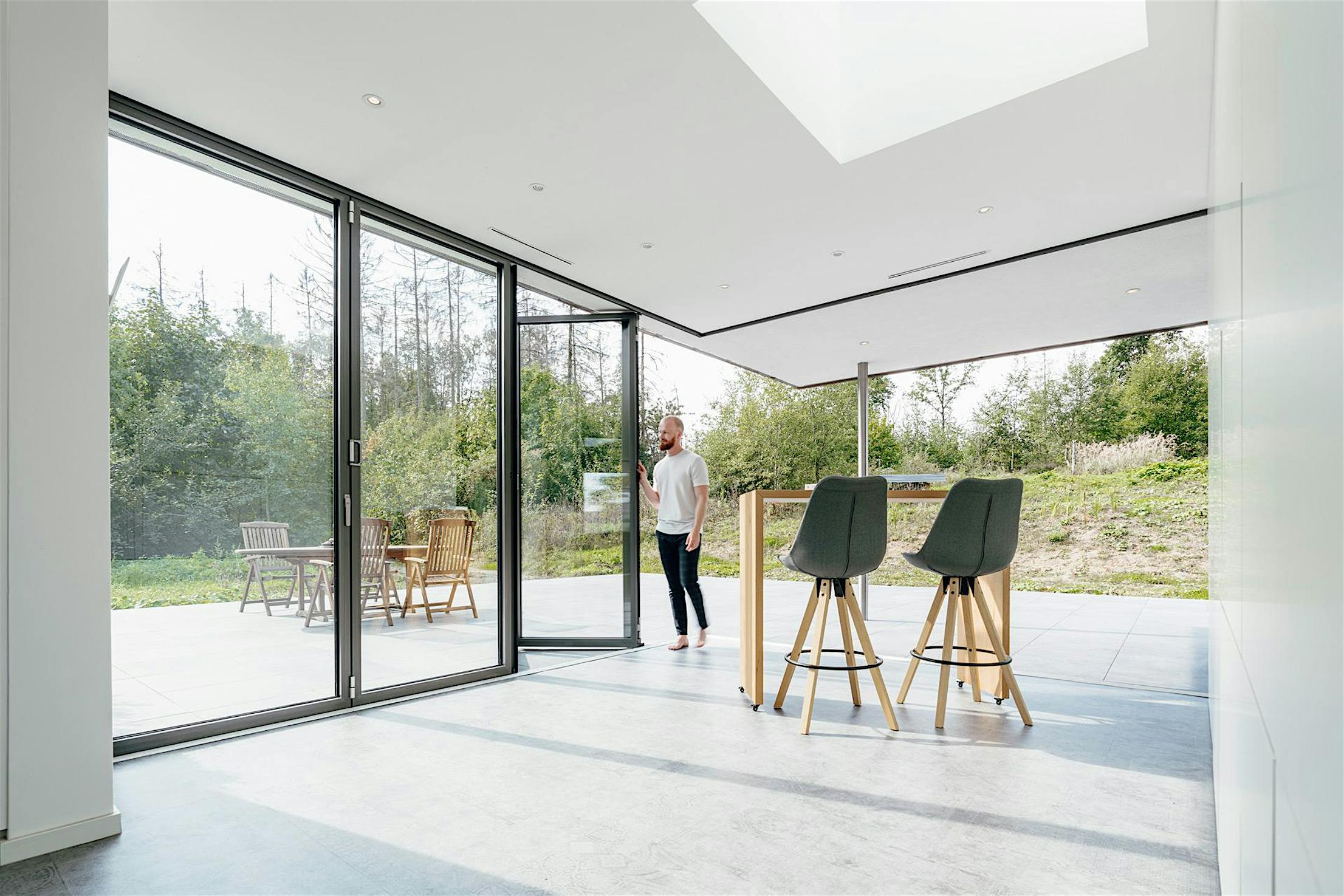 Person standing at open four-panel bifold glass doors, leading from a modern interior to a patio with outdoor furniture and greenery.