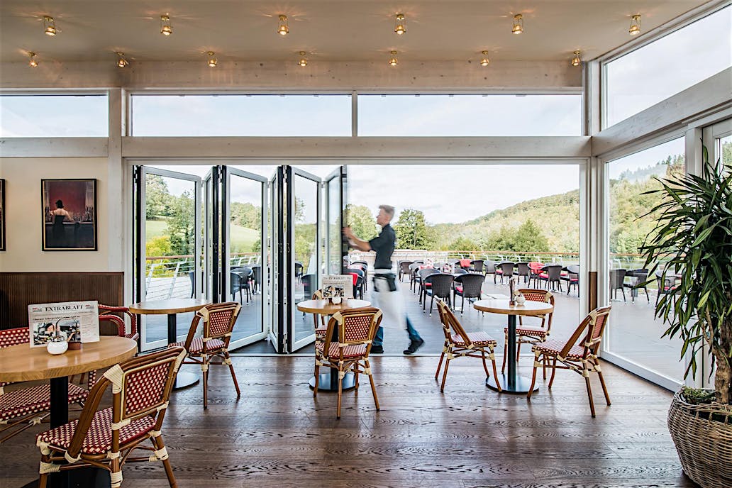 A server walks through commercial glass doors inside a bright café with wooden floors and wicker chairs, leading to an outdoor seating area.