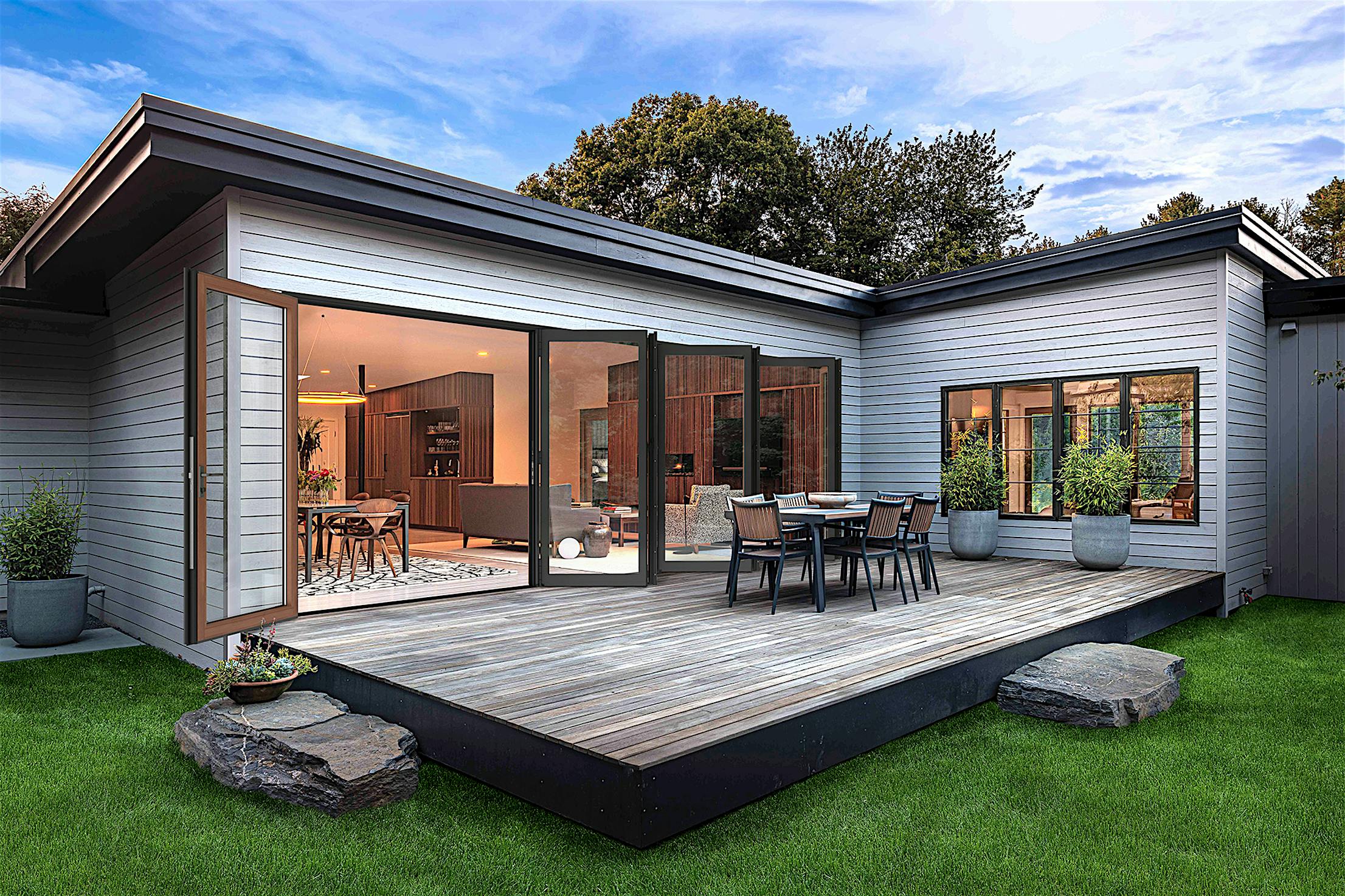 Modern house with large aluminum clad bifold glass doors opens to a wooden deck featuring an outdoor dining set, surrounded by green grass and potted plants.