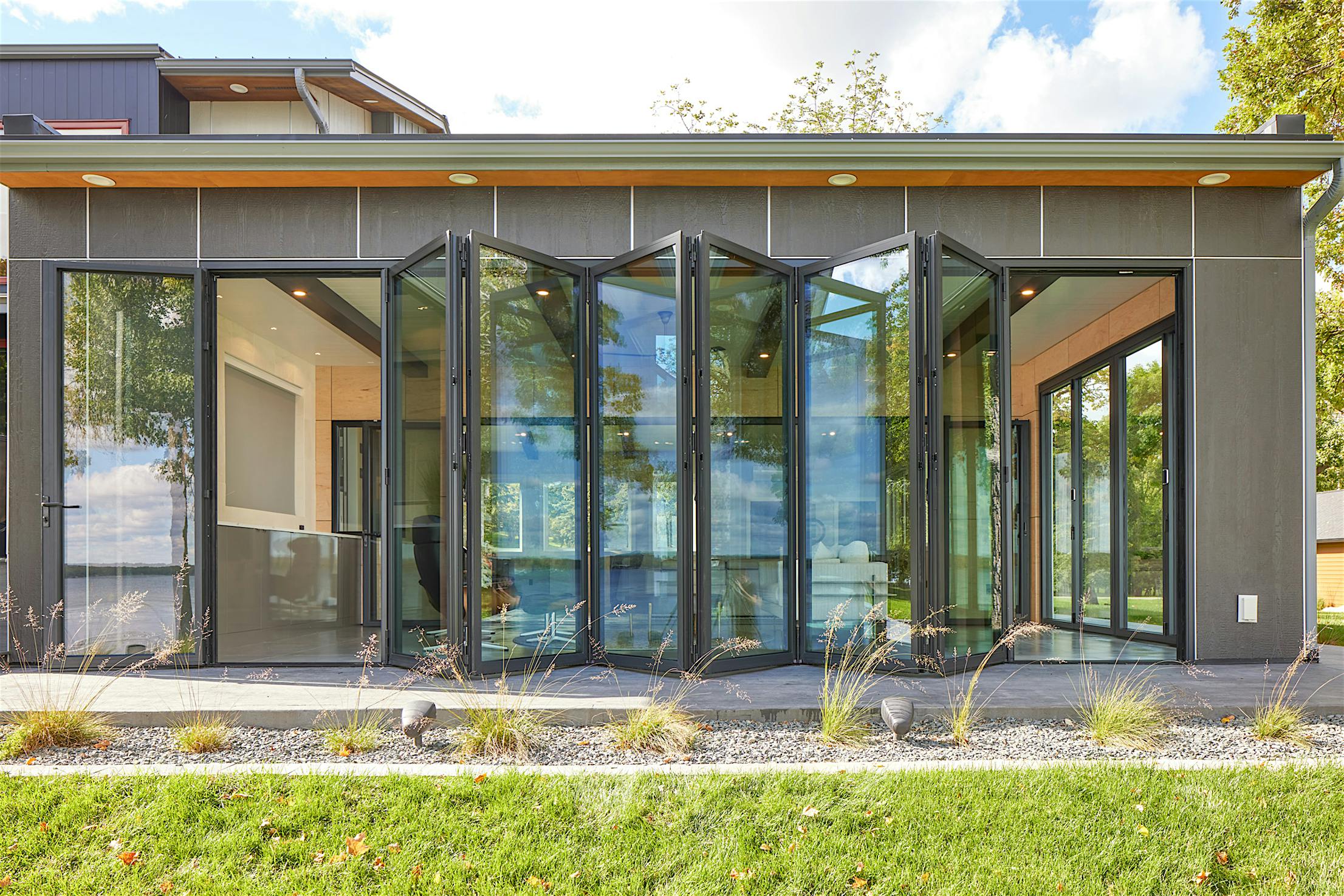 Modern building with large glass folding doors partially open, revealing an interior with wood accents and views of trees and grass outside.