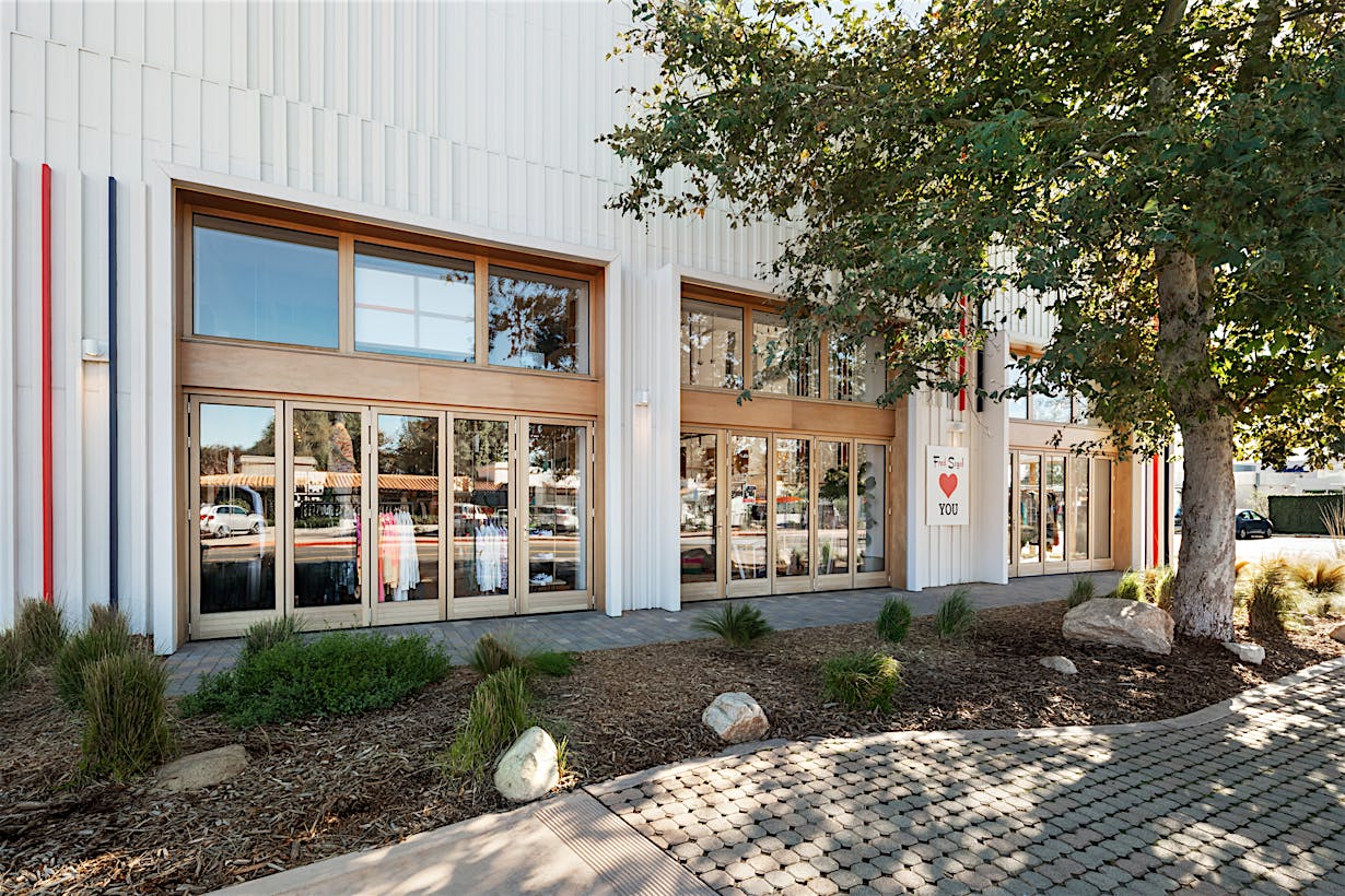 Exterior view of a modern commercial retail store with expansive glass doors and windows, surrounded by landscaping with trees and rocks, under a clear sky.