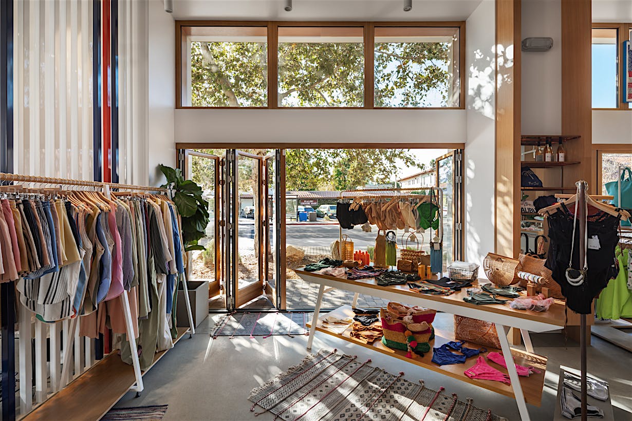 A brightly retail store features clothes on racks and accessories on tables, enhanced by accordion glass doors that offer a seamless view of trees through large windows and an open door.