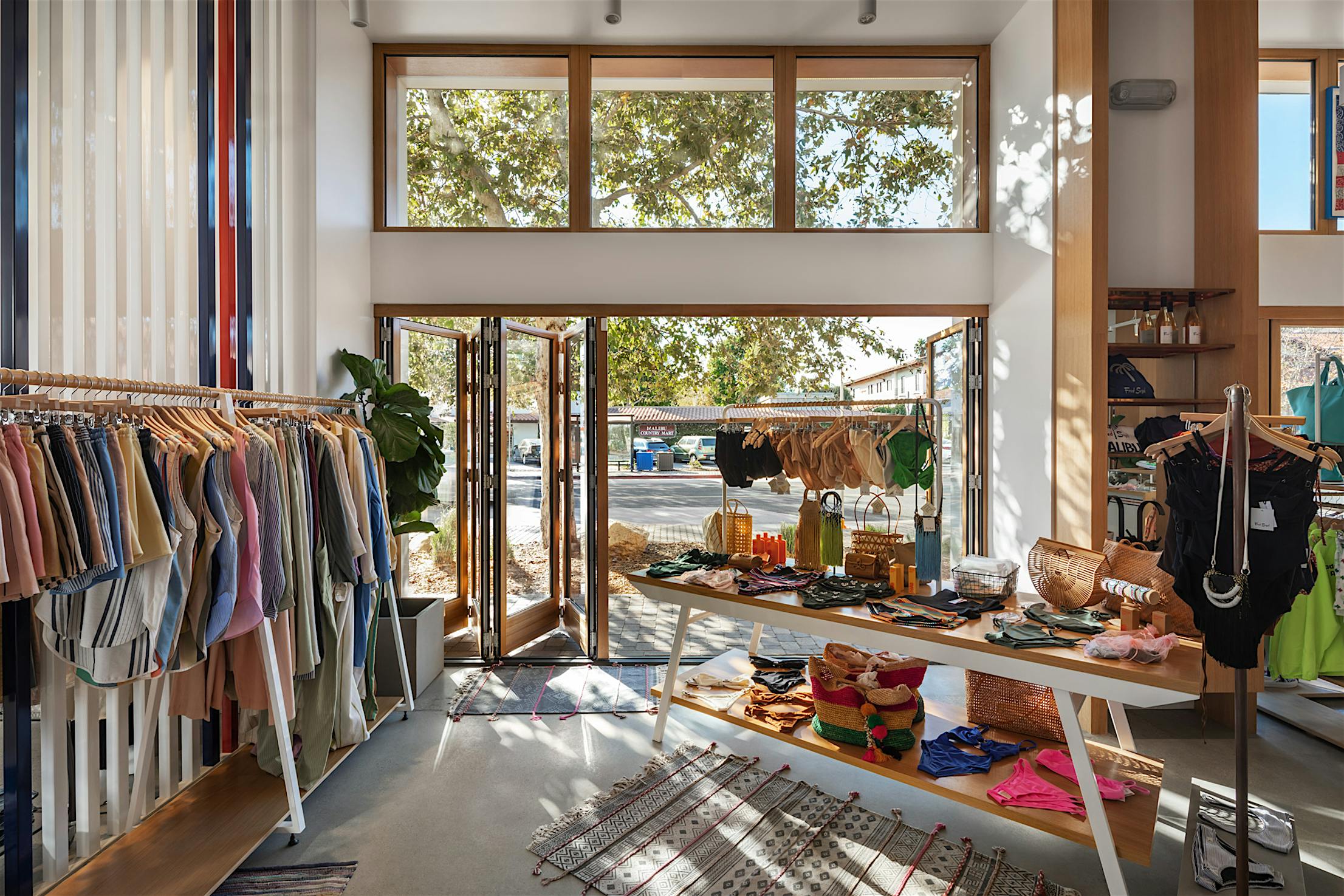 A brightly retail store features clothes on racks and accessories on tables, enhanced by accordion glass doors that offer a seamless view of trees through large windows and an open door.