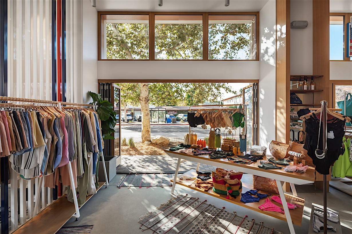 Retail store interior features racks of colorful garments and a central table displaying accessories. Large folding glass doors offer an expansive outdoor view, allowing natural light to stream in, creating a bright and inviting atmosphere.