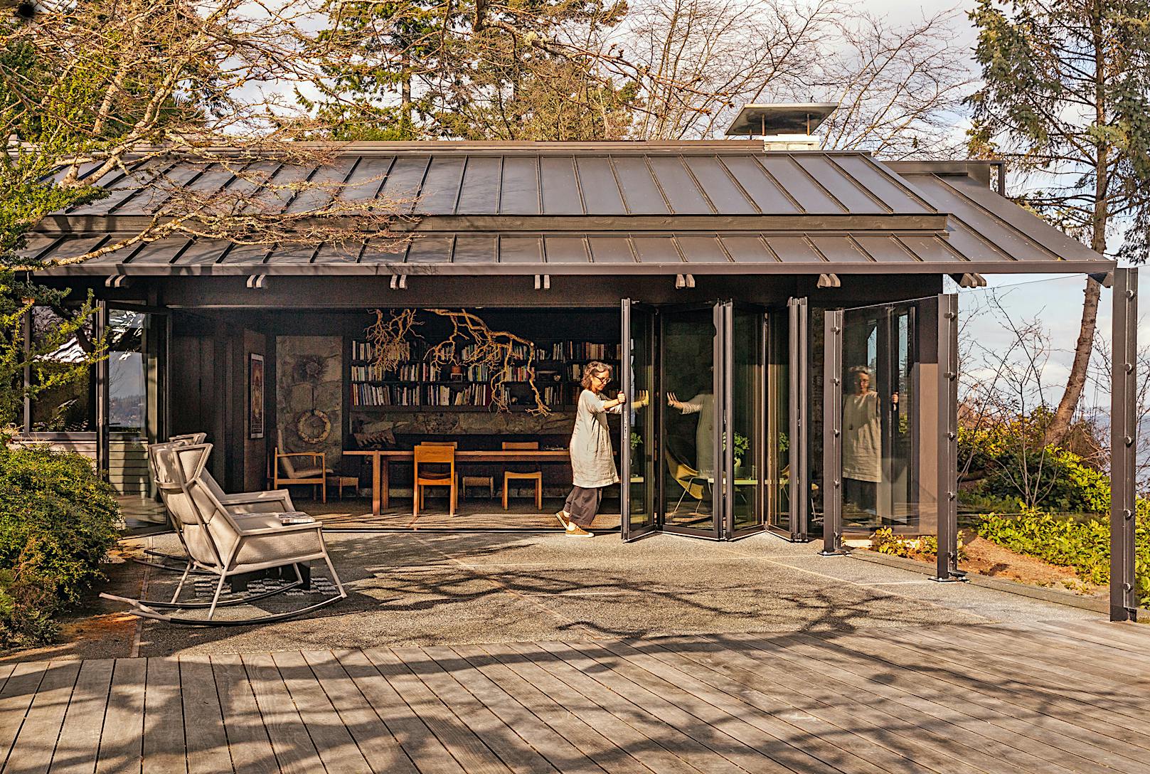 A person opens bifold glass doors of a modern building with a metal roof and outdoor seating. 