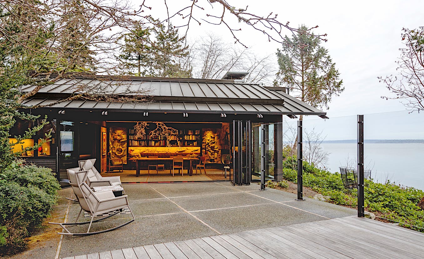 An open wooden ADU with bifold glass walls that seamlessly blend into the covered patio featuring two rocking chairs, overlooking a body of water and surrounded by trees. 