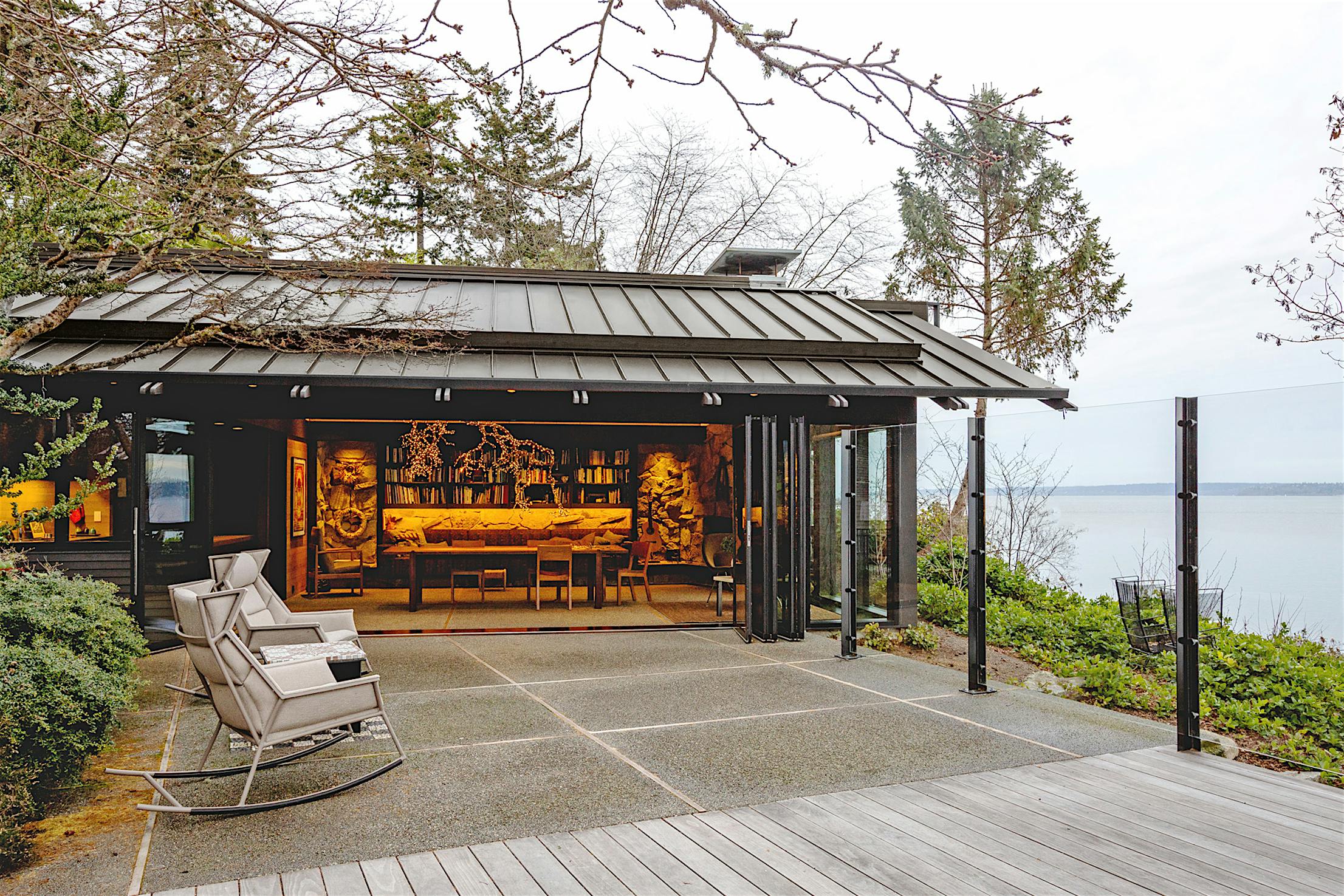 An open wooden ADU with bifold glass walls that seamlessly blend into the covered patio featuring two rocking chairs, overlooking a body of water and surrounded by trees.