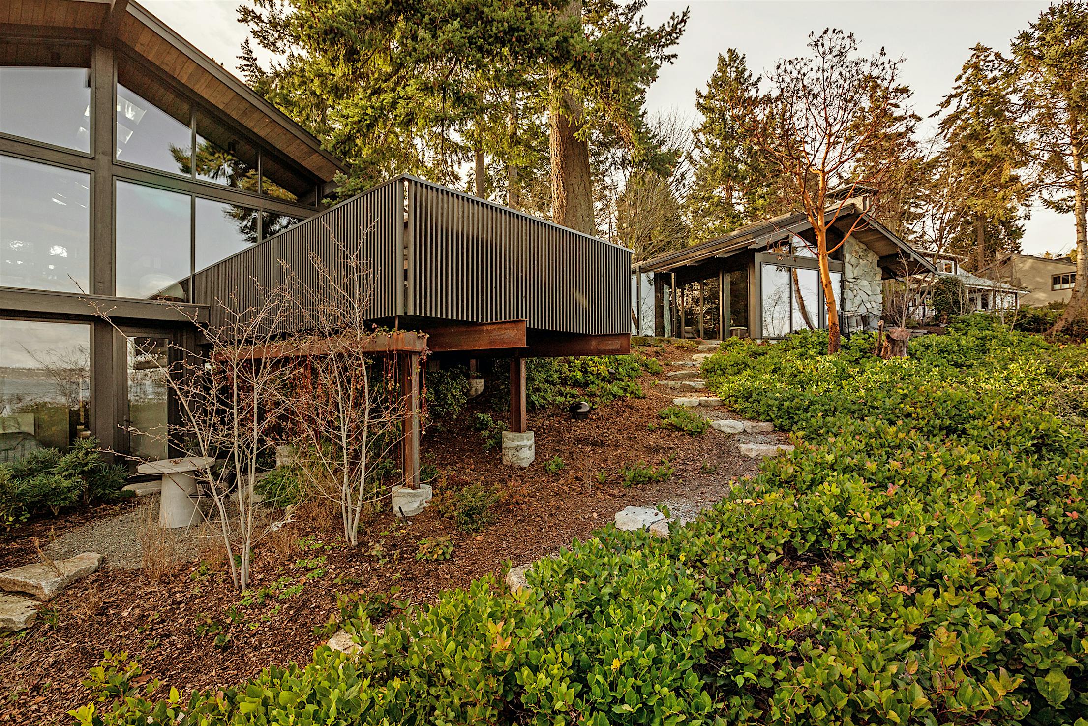 Modern house with large windows and glass walls, surrounded by greenery and trees.