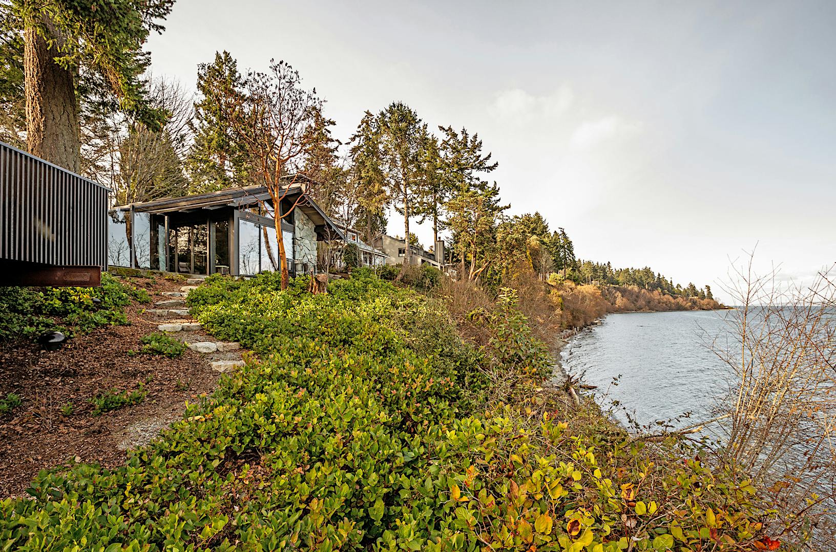 A house with large windows and glass doors is situated on a hillside overlooking a body of water.