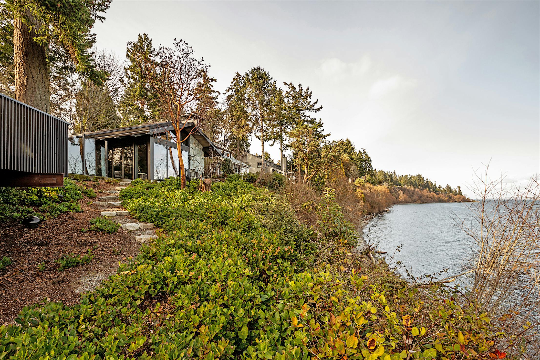 A house with large windows and glass doors is situated on a hillside overlooking a body of water.