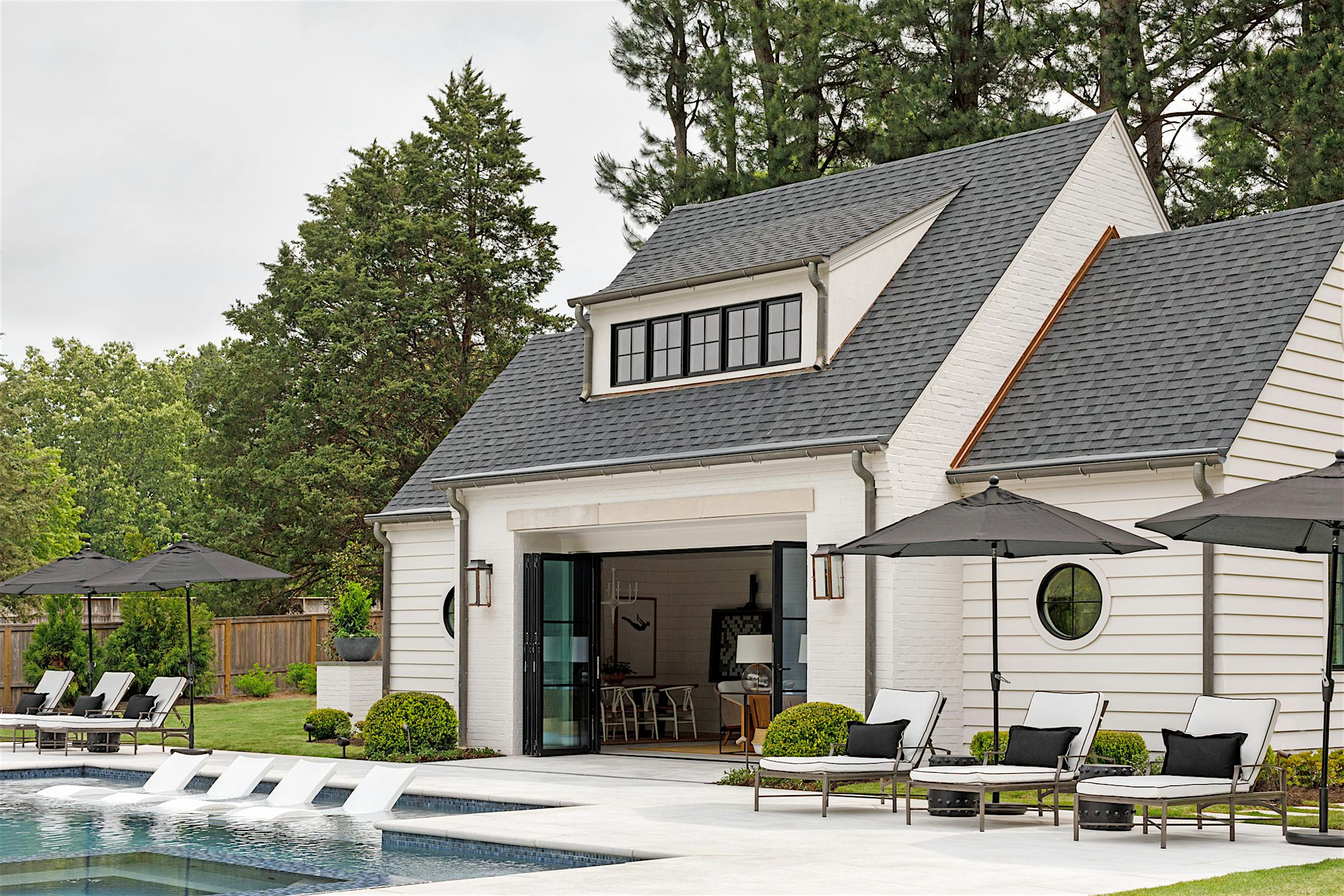 Outdoor pool area with lounge chairs and umbrellas in front of a white house featuring large windows and folding glass walls leading to an indoor room.