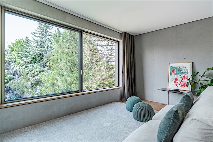 Modern living room with large sliding windows, tree views, gray sofa with blue pillows, and framed artwork.