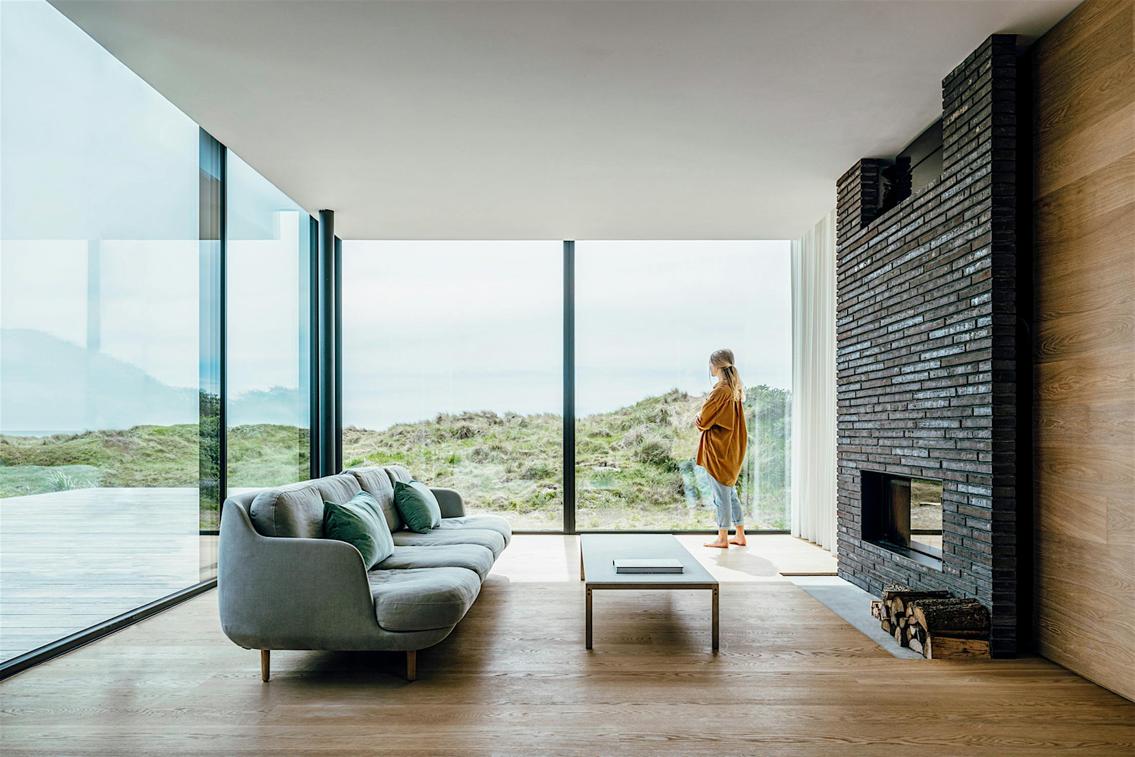 Person standing by large sliding glass doors in a modern living room with minimalist furniture, overlooking seaside dunes.