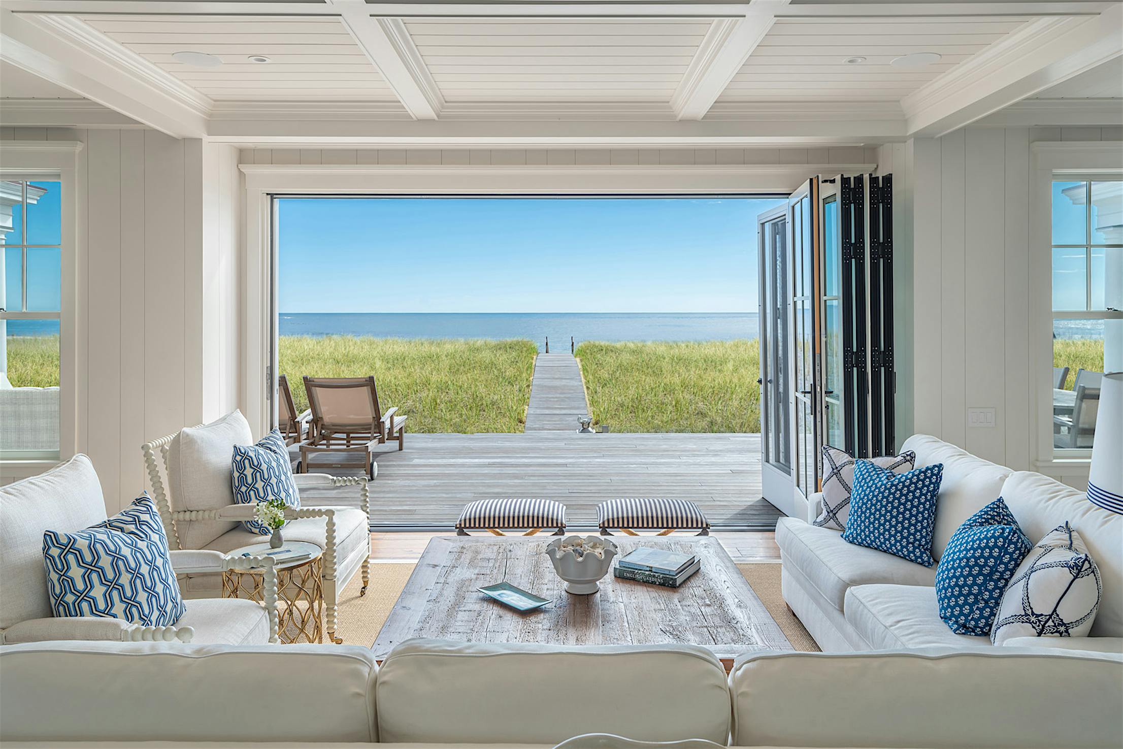 Modern living room features a large coffee table, sofas, and armchairs, all facing the ocean view through aluminum clad retractable glass doors.