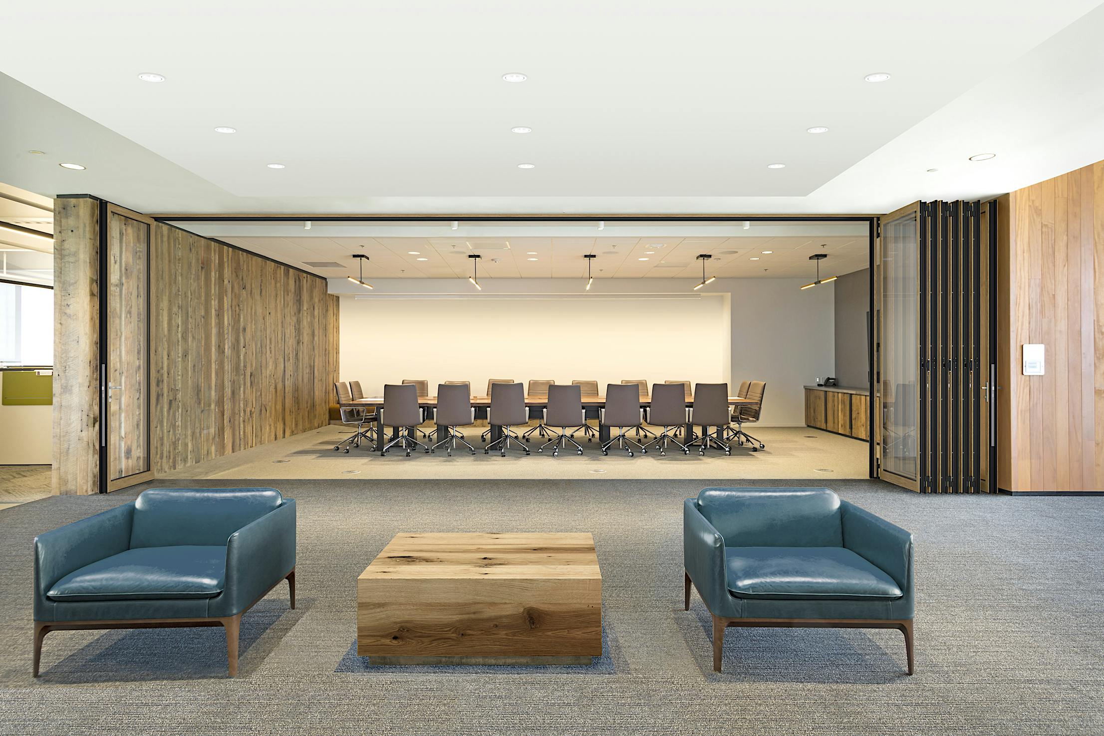 Modern office lobby with two blue armchairs and a wooden coffee table, facing a conference room opened folding glass doors.