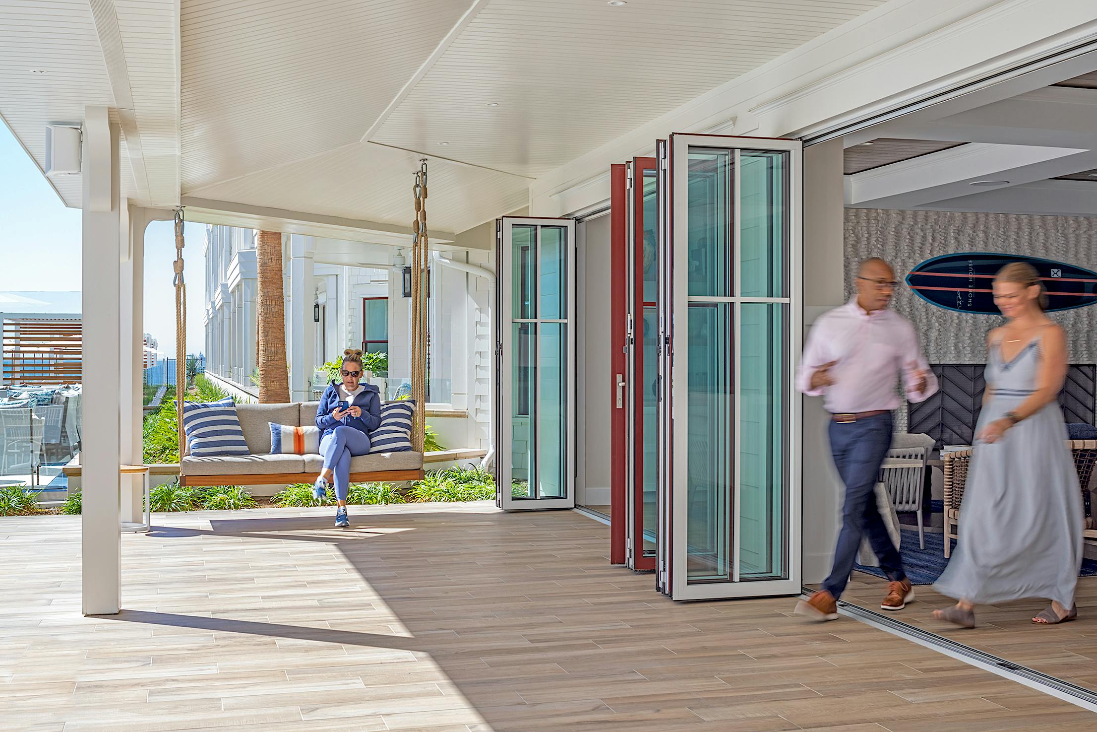 A man sits on a suspended outdoor bench while a man and a woman walk through an open folding glass door into a bright, modern hotel lobby area.
