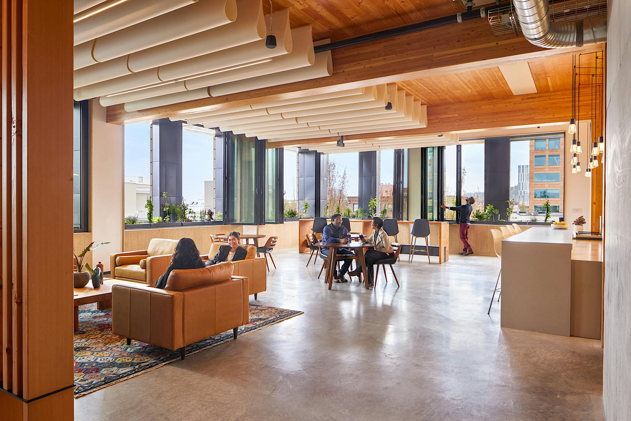 Modern office lounge with sofas, a table, wooden ceiling, indoor plants, and large folding windows for ample natural light.