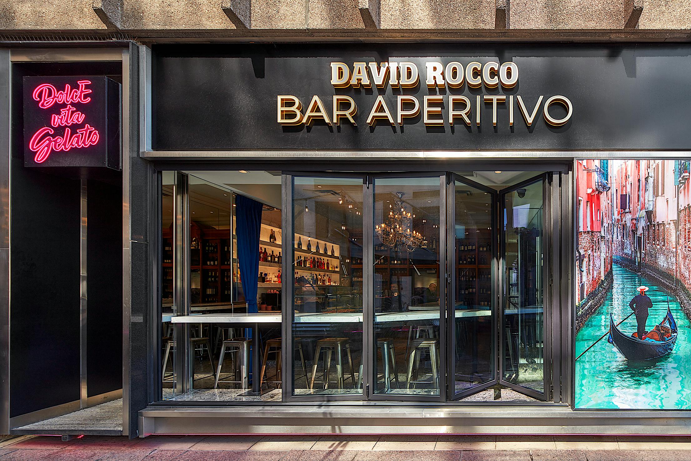 Exterior of 'David Rocco Bar Aperitivo' featuring folding glass doors, a neon 'Dolce Vita Gelato' sign, and a gondola photo, perfect for an authentic Italian experience.