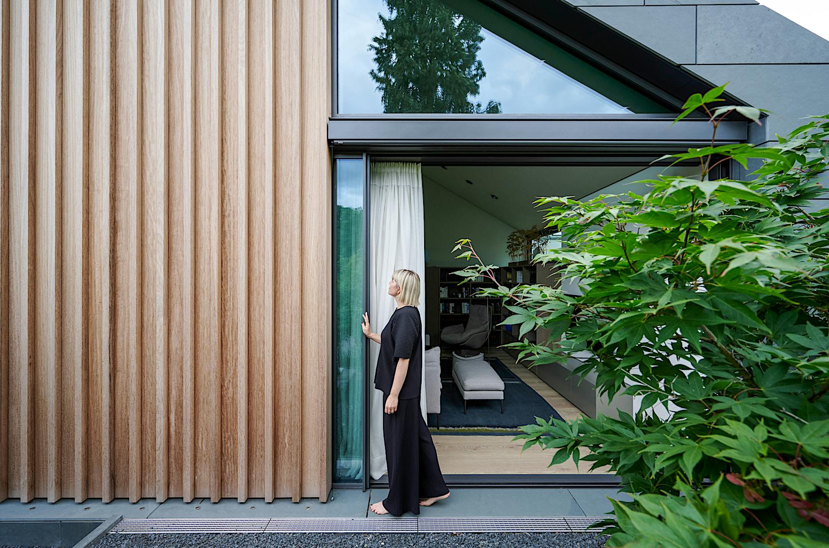 A woman is standing outside of a modern house with sliding glass walls