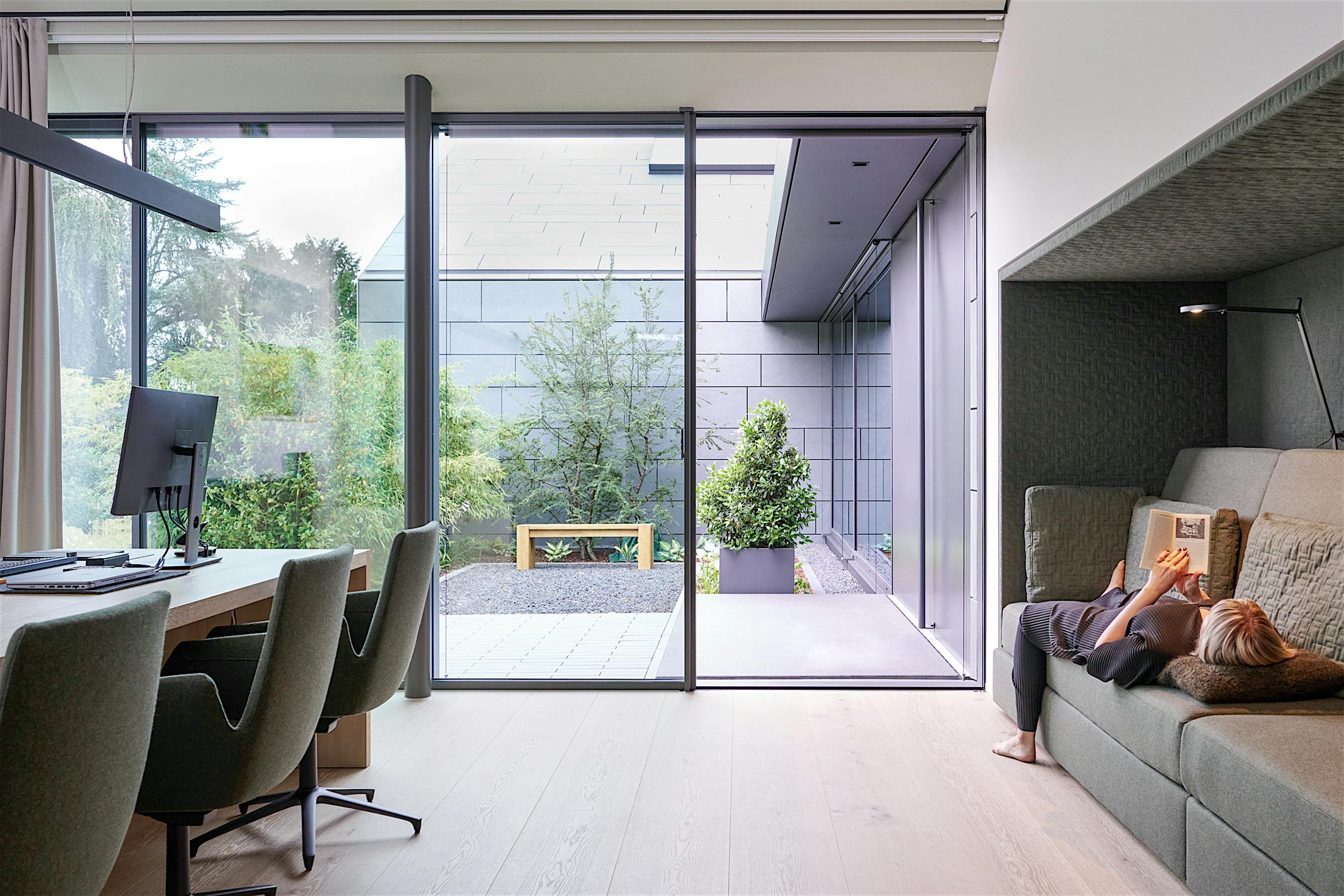 A woman is reading a book on a couch in a home office with sliding glass walls