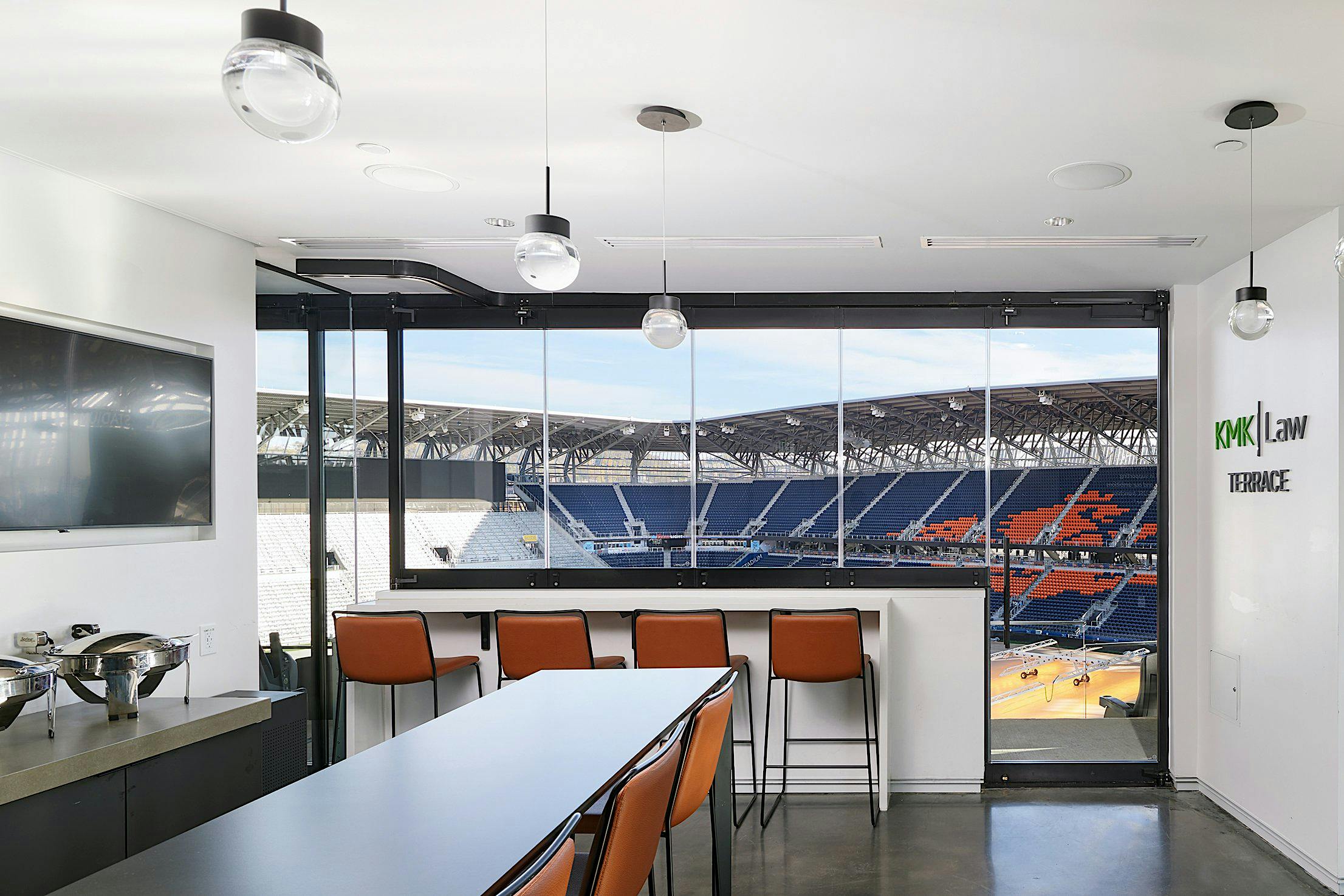 Modern stadium suite featuring commercial sliding windows with integrated swing door, bar seating overlooking the field, and 'KMK Law Terrace'—ideal for VIP and corporate events.