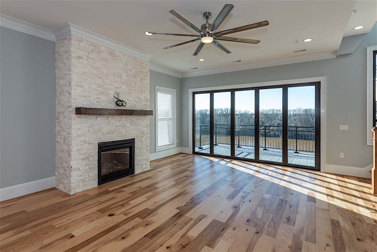 A modern living room with a wooden floor, a stone fireplace, a ceiling fan, and large glass doors that open to a balcony with an outdoor view.