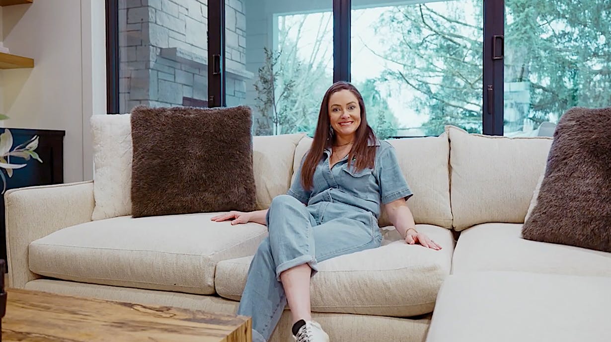 Molly Mesnick in casual clothing sits in a modern living room featuring large bifold glass walls, which open up to a scenic view.