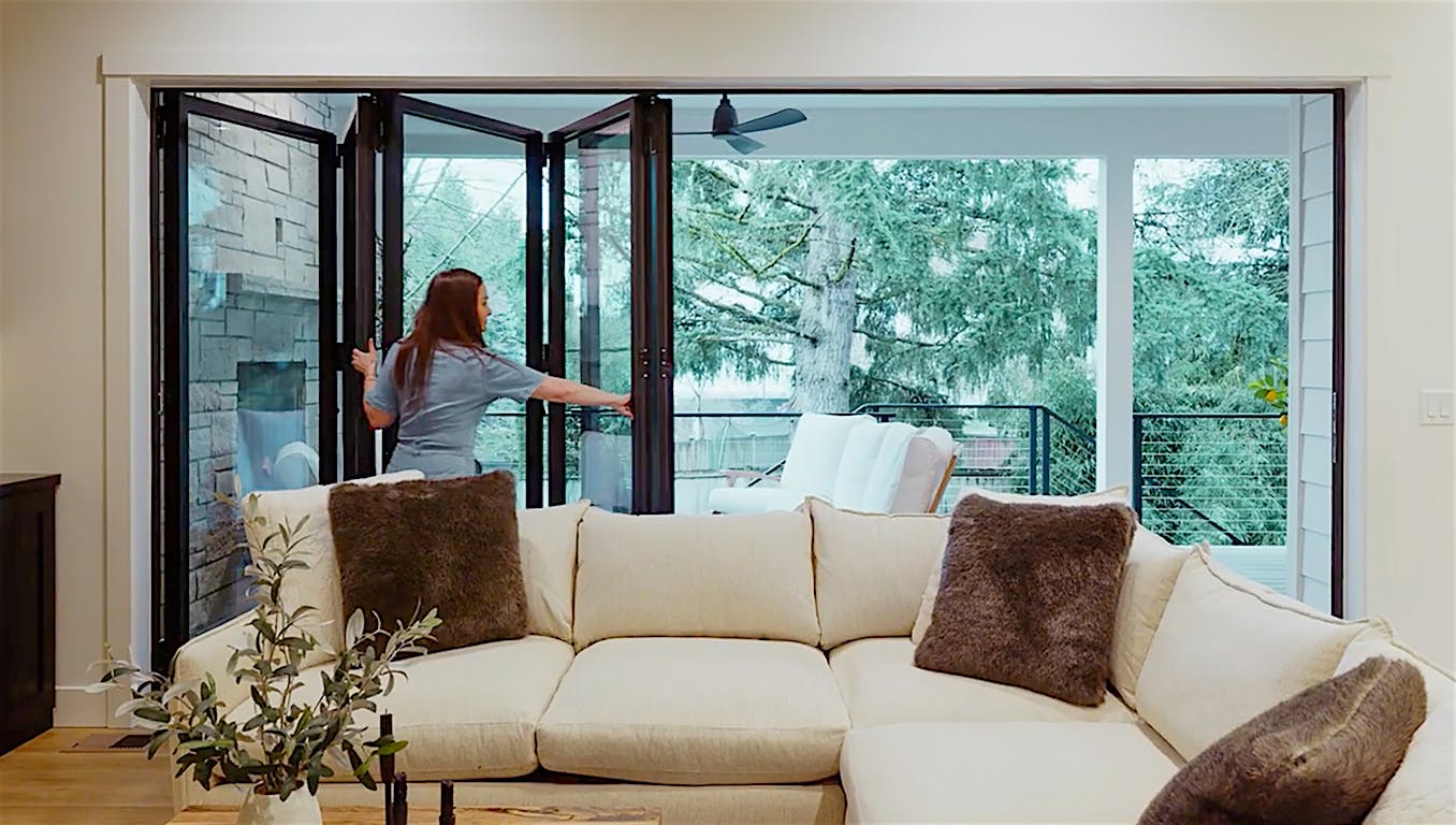 Molly Mesnick is opening a set of bifold doors in a modern living room leading to an outdoor seating area with greenery visible beyond.