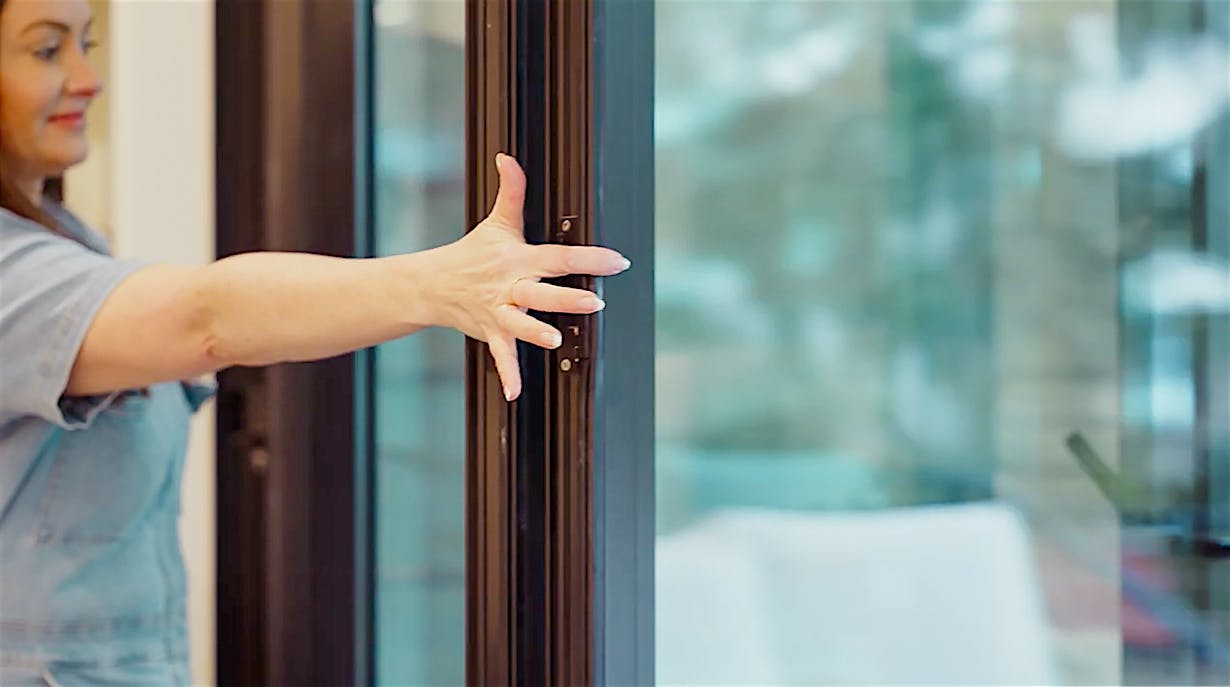 Molly Mesnick pushes a folding glass door closed with their hand, seamlessly connecting the sleek interior with the blurred outdoor view, framed by elegant glass walls.