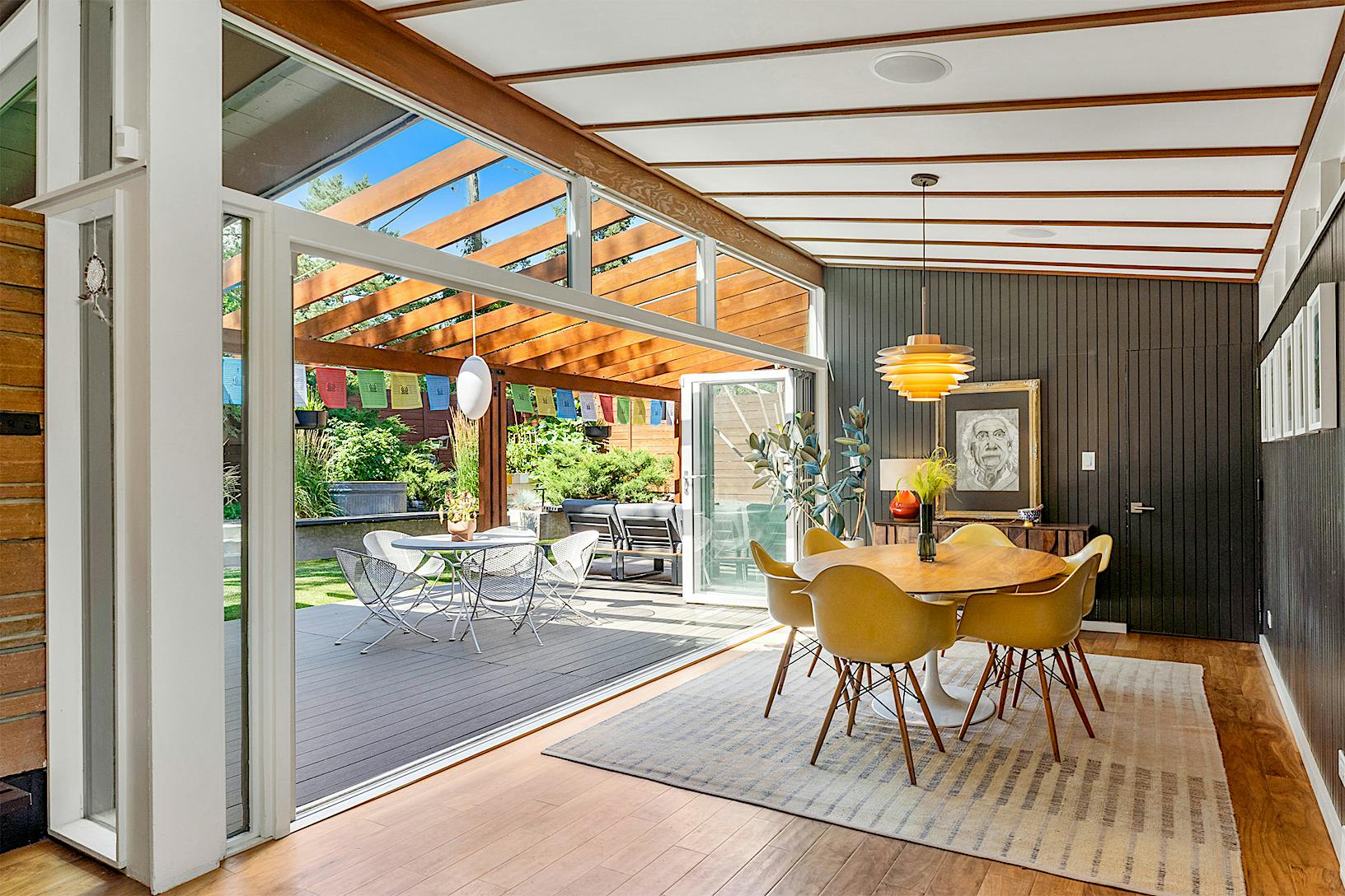 A modern dining area with wooden ceiling beams and a table with yellow chairs opens to an outdoor patio with seating through folding glass doors. 