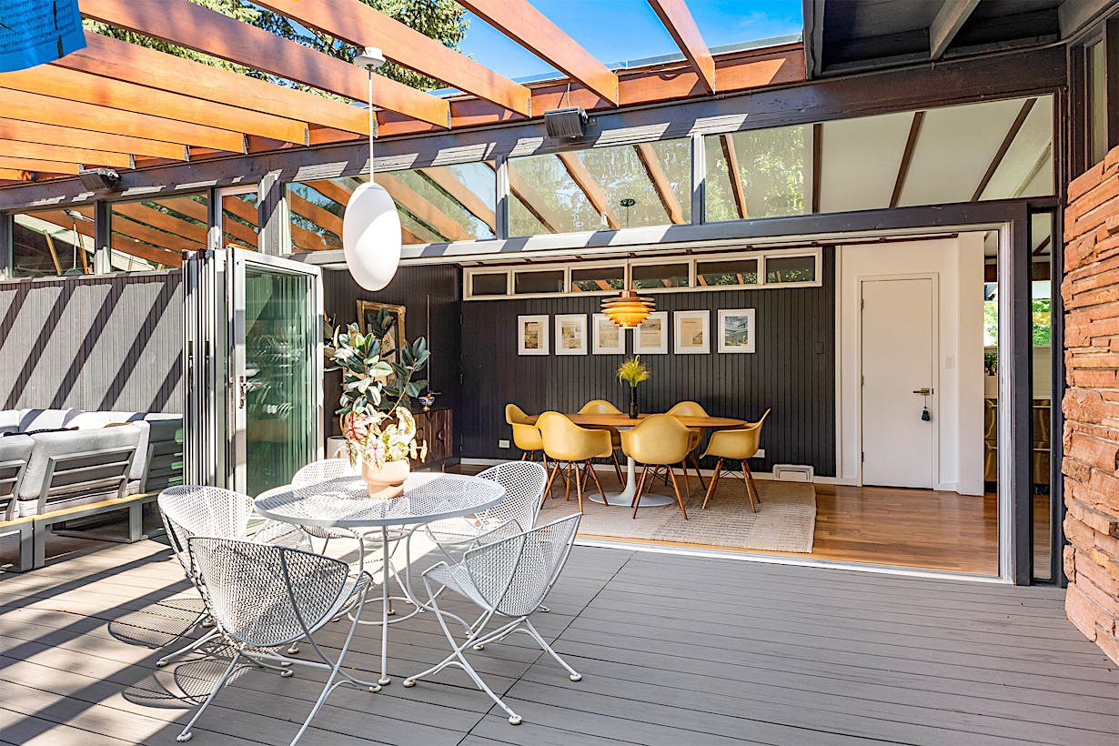 Patio with white mesh furniture and a hanging light overlooks an indoor dining area with yellow chairs and a wooden table, separated by large folding glass patio doors.