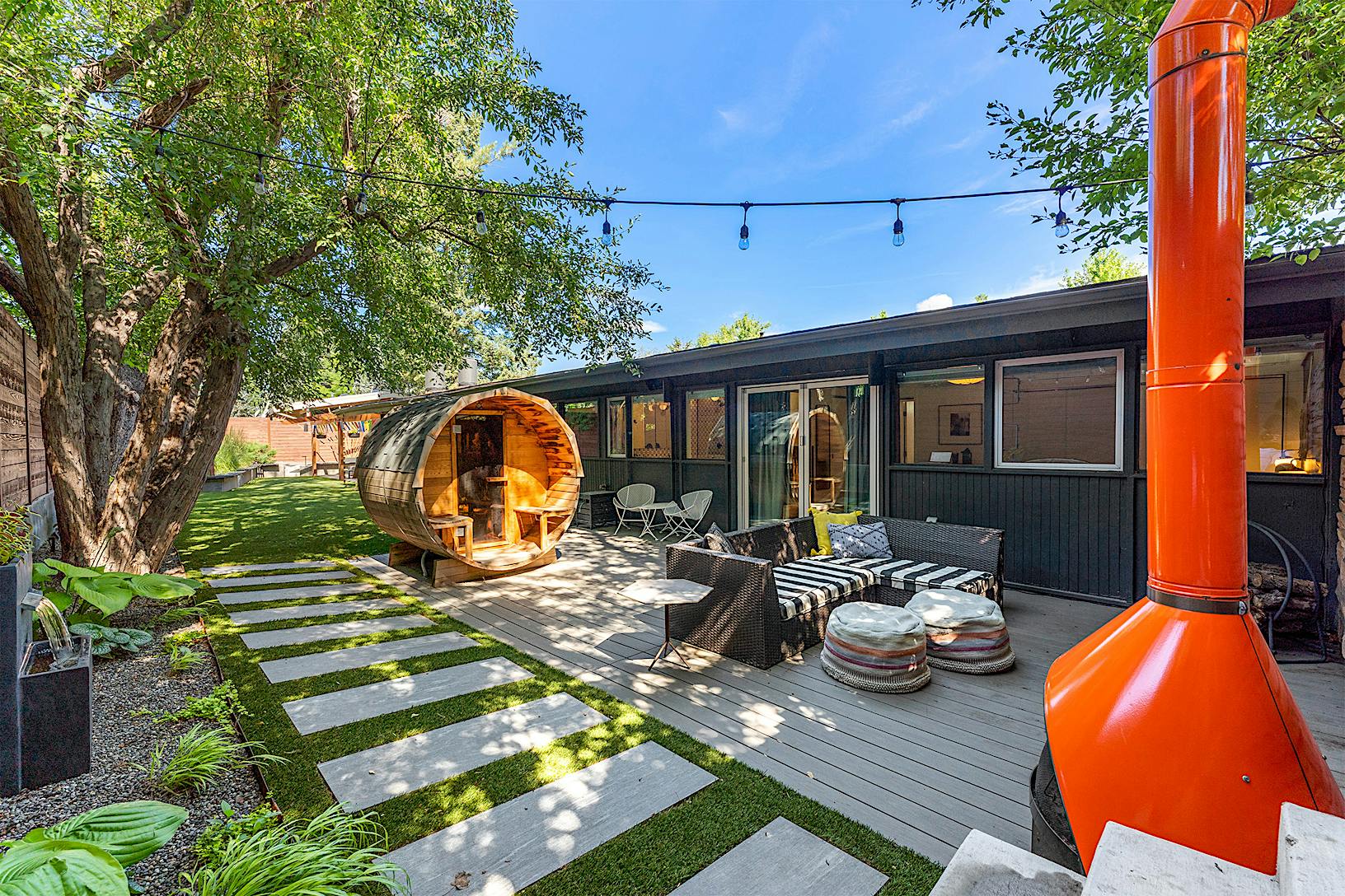A modern backyard featuring a wooden deck with seating, a unique cylindrical wooden sauna, glass doors leading to the house.