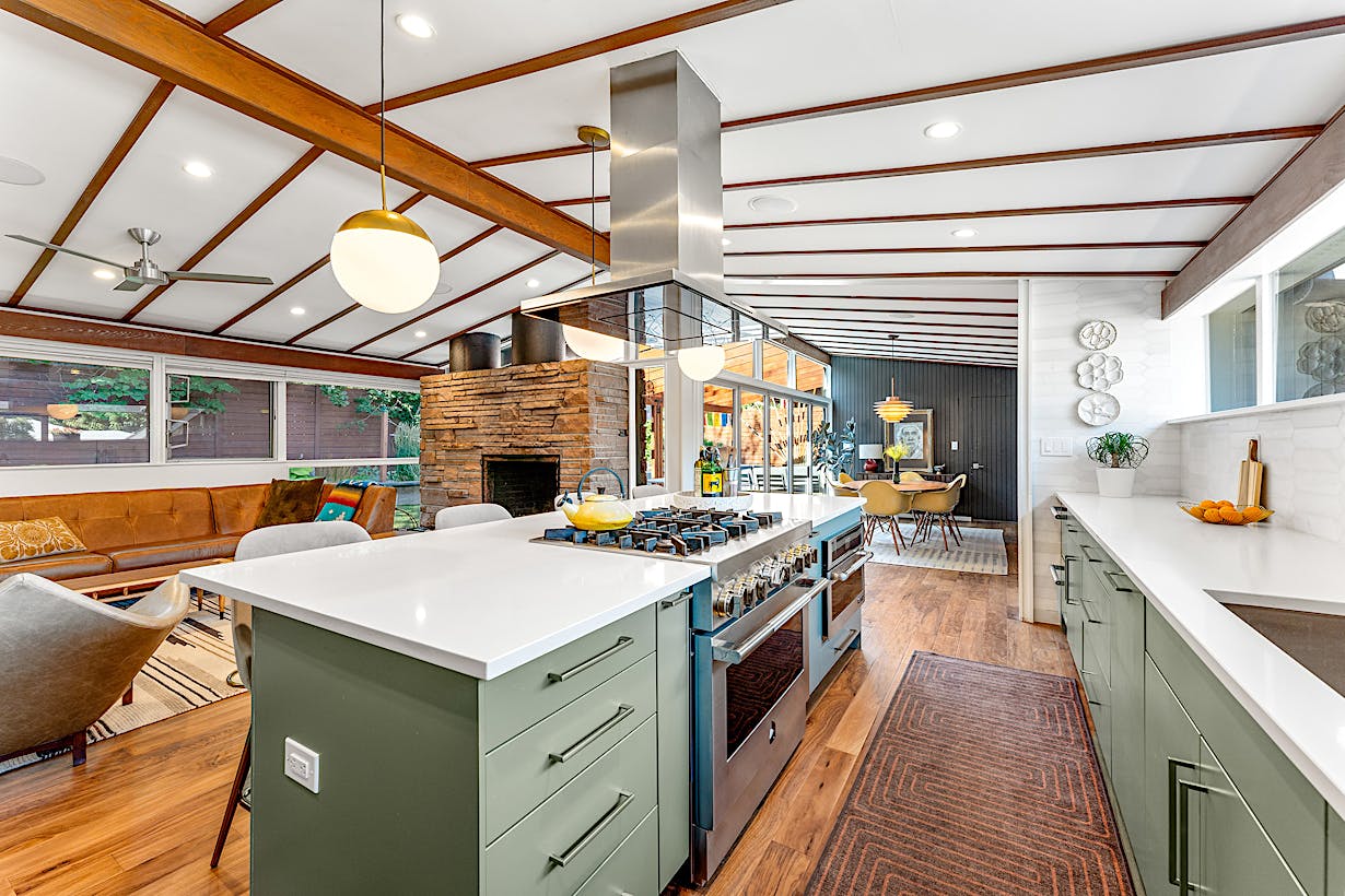 Modern open-plan kitchen and living area featuring a large island with a stovetop and hood, green cabinetry, a stone fireplace, wood flooring, and folding glass doors connecting to the patio area