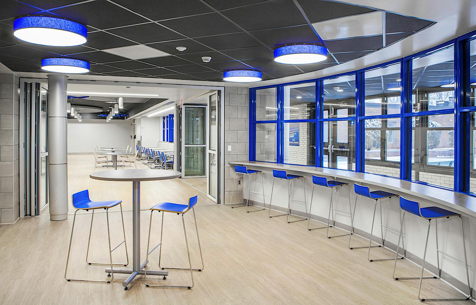 Modern room with high-top tables and chairs, a long counter with stools by large blue-framed windows