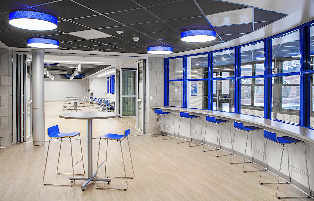 Modern room with high-top tables and chairs, a long counter with stools by large blue-framed windows