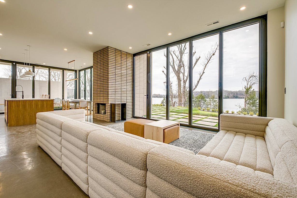 A modern living room with a large sectional sofa, a brick fireplace, floor-to-ceiling accordion glass doors offering a stunning view of trees and a body of water.