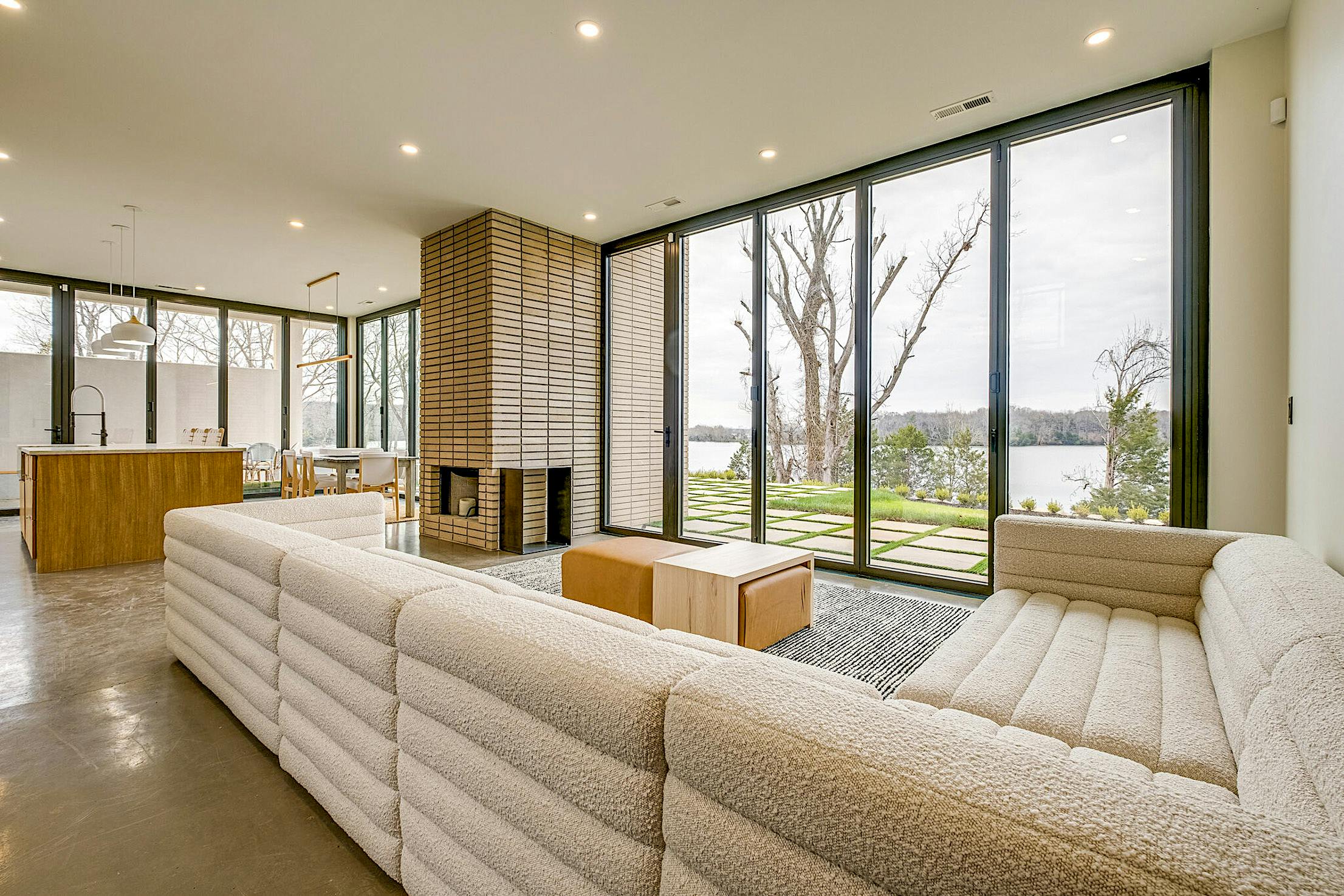 A modern living room with a large sectional sofa, a brick fireplace, floor-to-ceiling accordion glass doors offering a stunning view of trees and a body of water.