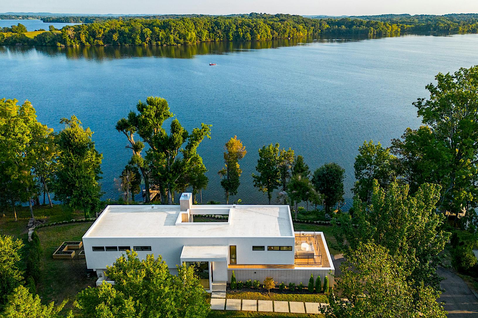Aerial view of a modern, white flat-roofed house surrounded by lush trees, located near the edge of a large lake.