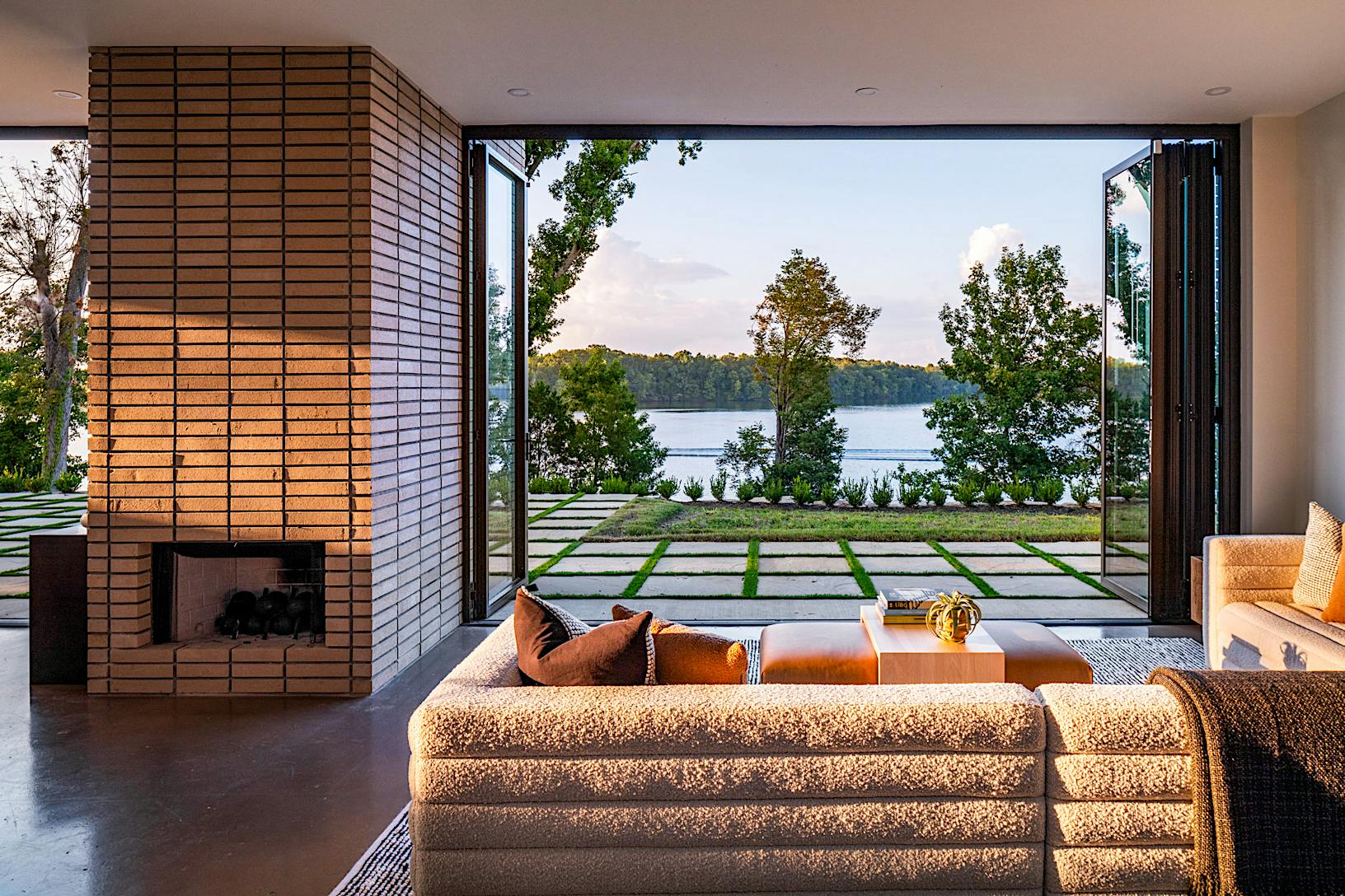 A modern living room with folding patio doors reveals a view of a lake and landscaped garden. 