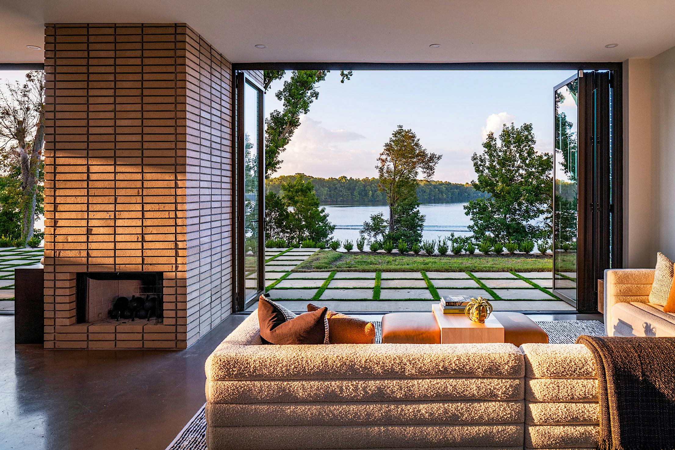 A modern living room with folding patio doors reveals a view of a lake and landscaped garden.
