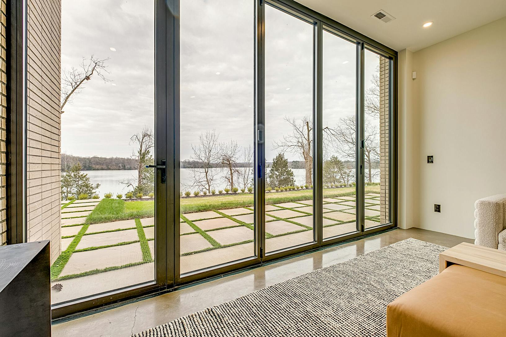 Modern living room with all glass walls overlooking a patio and lawn beside a lake.