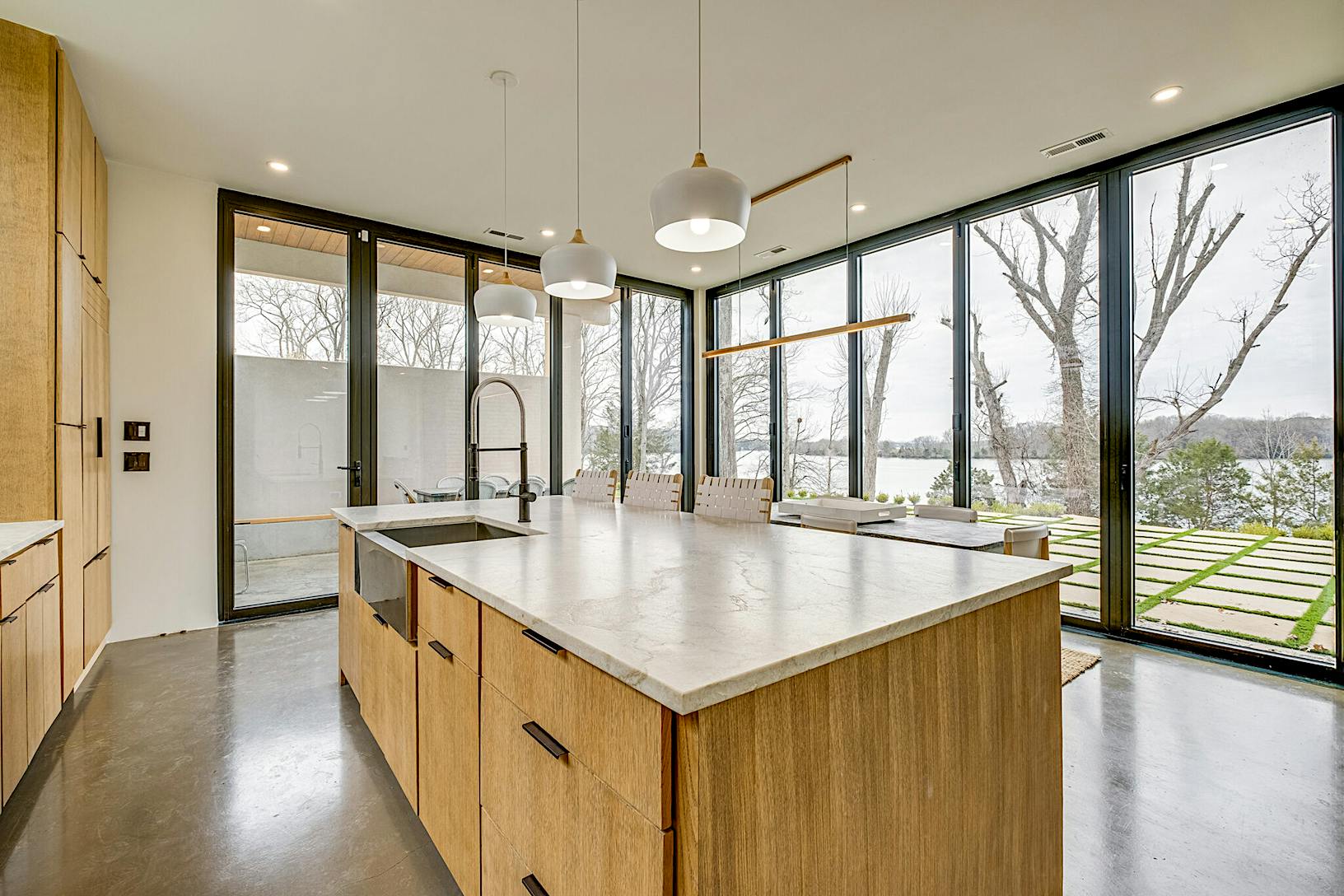 A modern kitchen featuring a large island with a stone countertop and large opening folding glass walls offering a view of the lake and trees in the background.